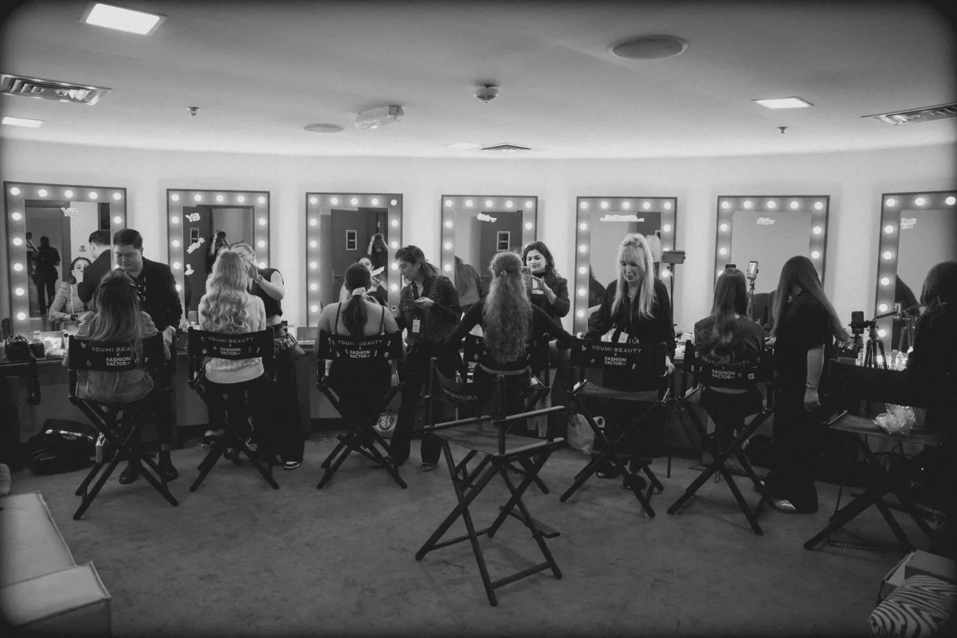 Backstage makeup room with illuminated vanity mirrors and stylists applying makeup to models seated in chairs at each station of a busy studio judge