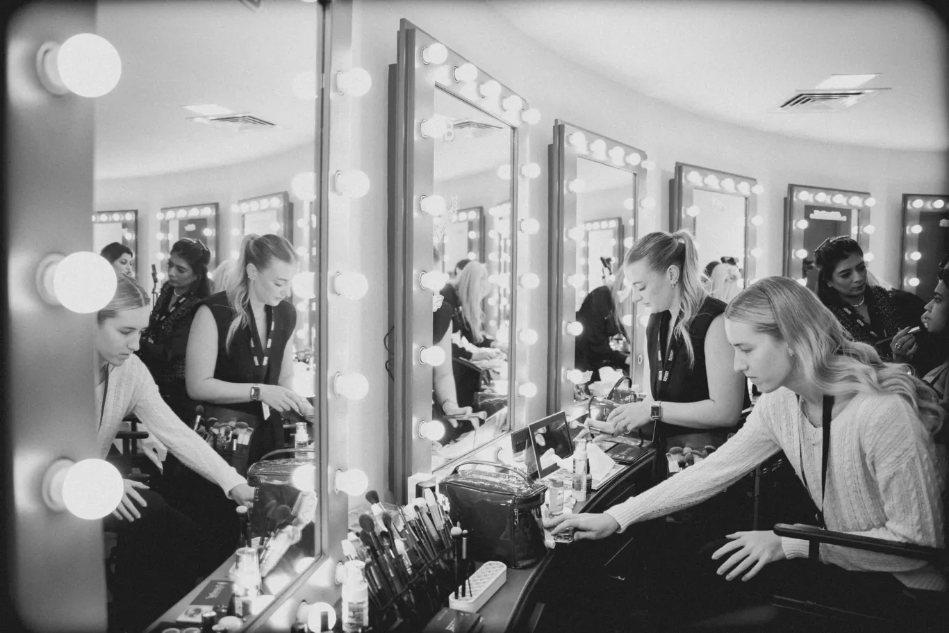 Backstage makeup station with illuminated vanity mirrors and several women applying makeup.