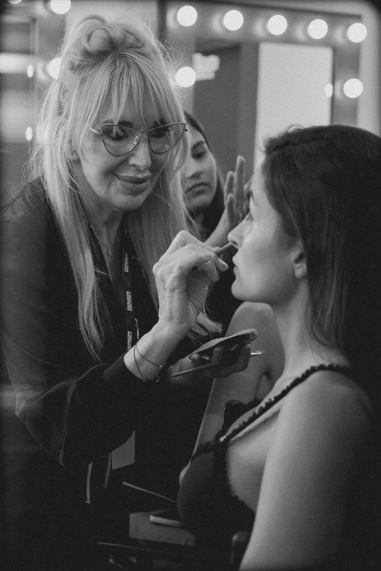 Makeup artist applies lipstick to a client in a backstage makeup area with bright vanity lights behind them