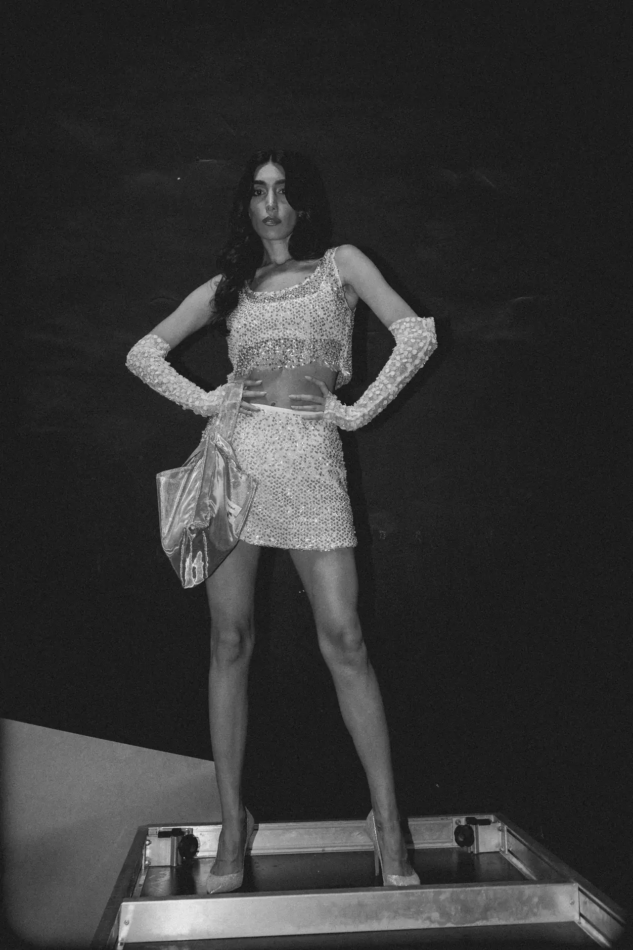 Woman in a sparkling costume posing on a stage-like platform during a fashion event, hands on hips, clutch bag at side.