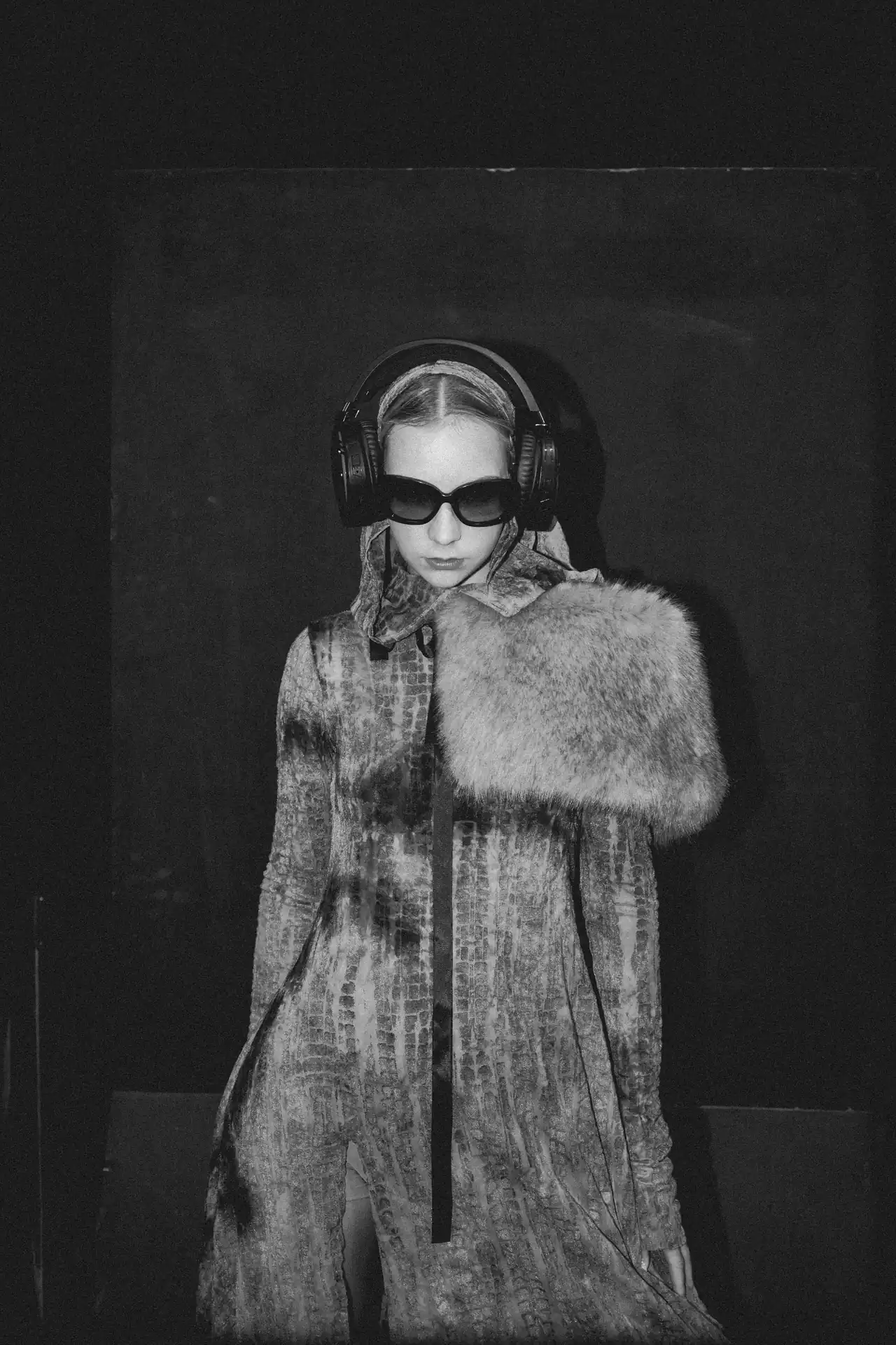 Person wearing oversized black sunglasses and large headphones, with a fur-trimmed coat in a dark studio.