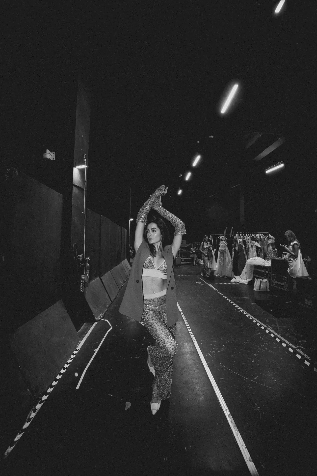 Backstage at a fashion show: a model poses in a glittery outfit with a coat, clothing racks visible in the background.