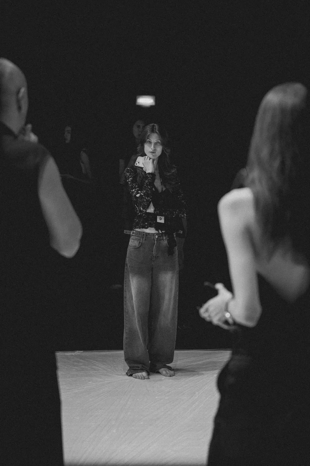 Woman standing barefoot on a stage, facing the camera, wearing a lace top and jeans, holding a smartphone near her chest with an audience blurred behind.