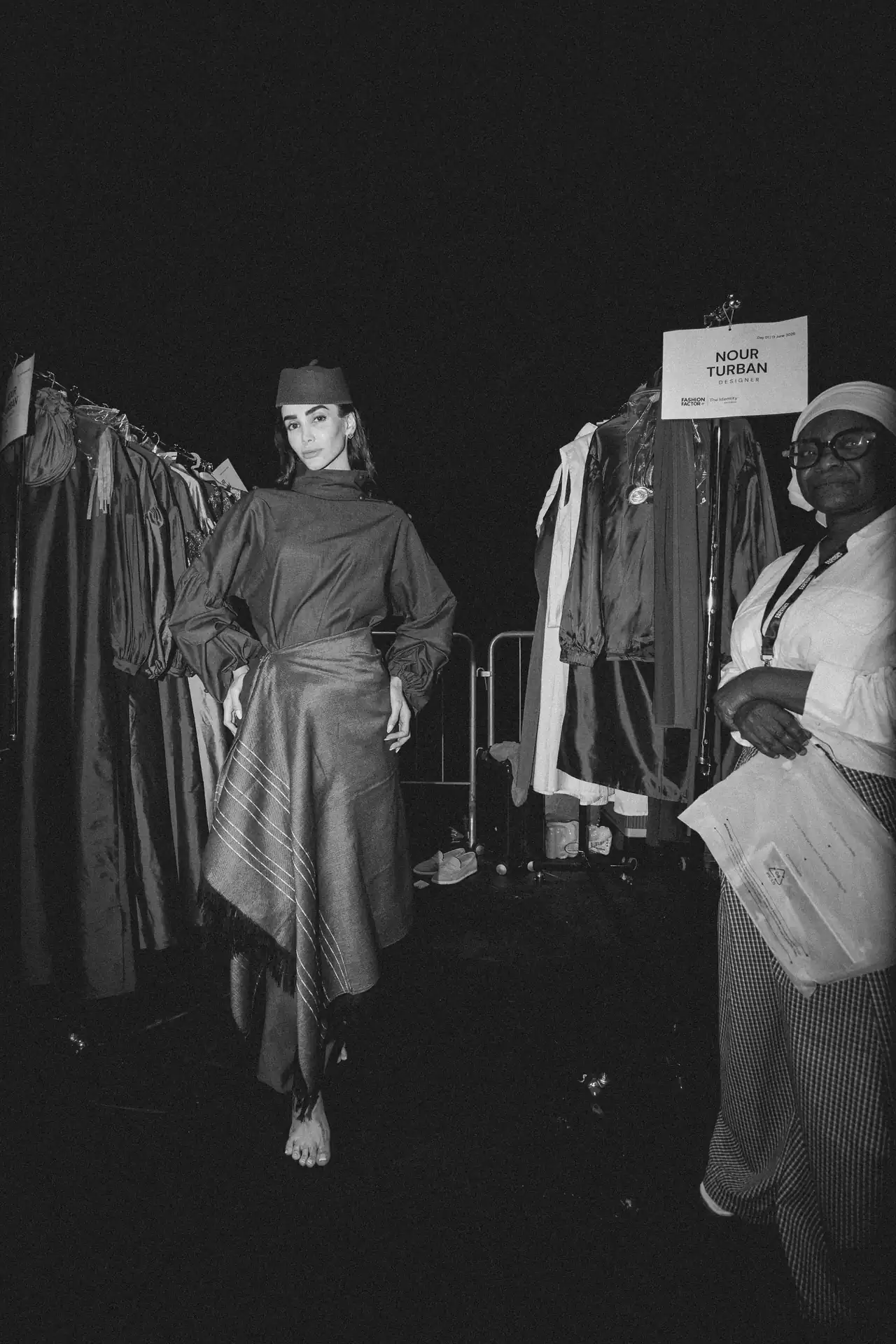 Model backstage on a fashion show runway wearing a structured, long dress with a wrapped waist and headpiece, barefoot on a dark floor with racks of clothing nearby.
