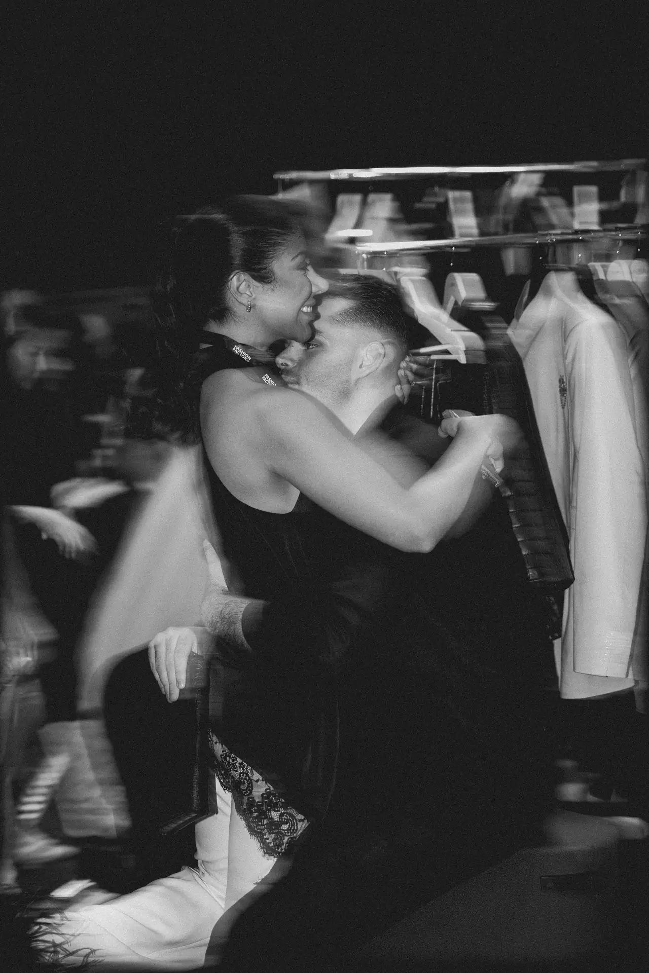 Two people embrace and dance closely in a dimly lit clothing store, with racks of shirts visible in the background.