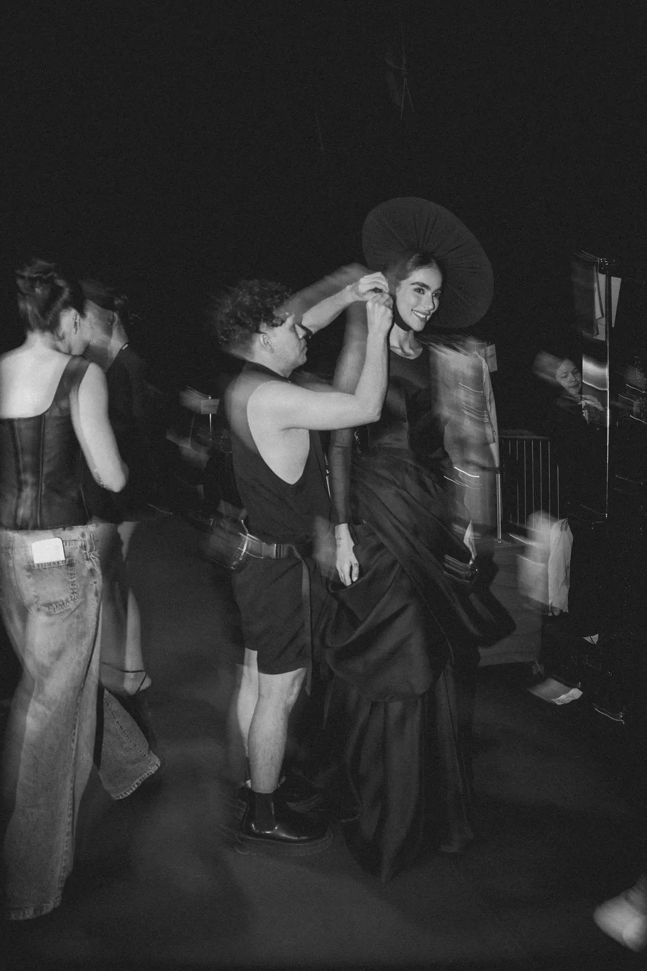 Backstage fashion moment: a man adjusts an oversized circular headpiece on a smiling model as others look on.