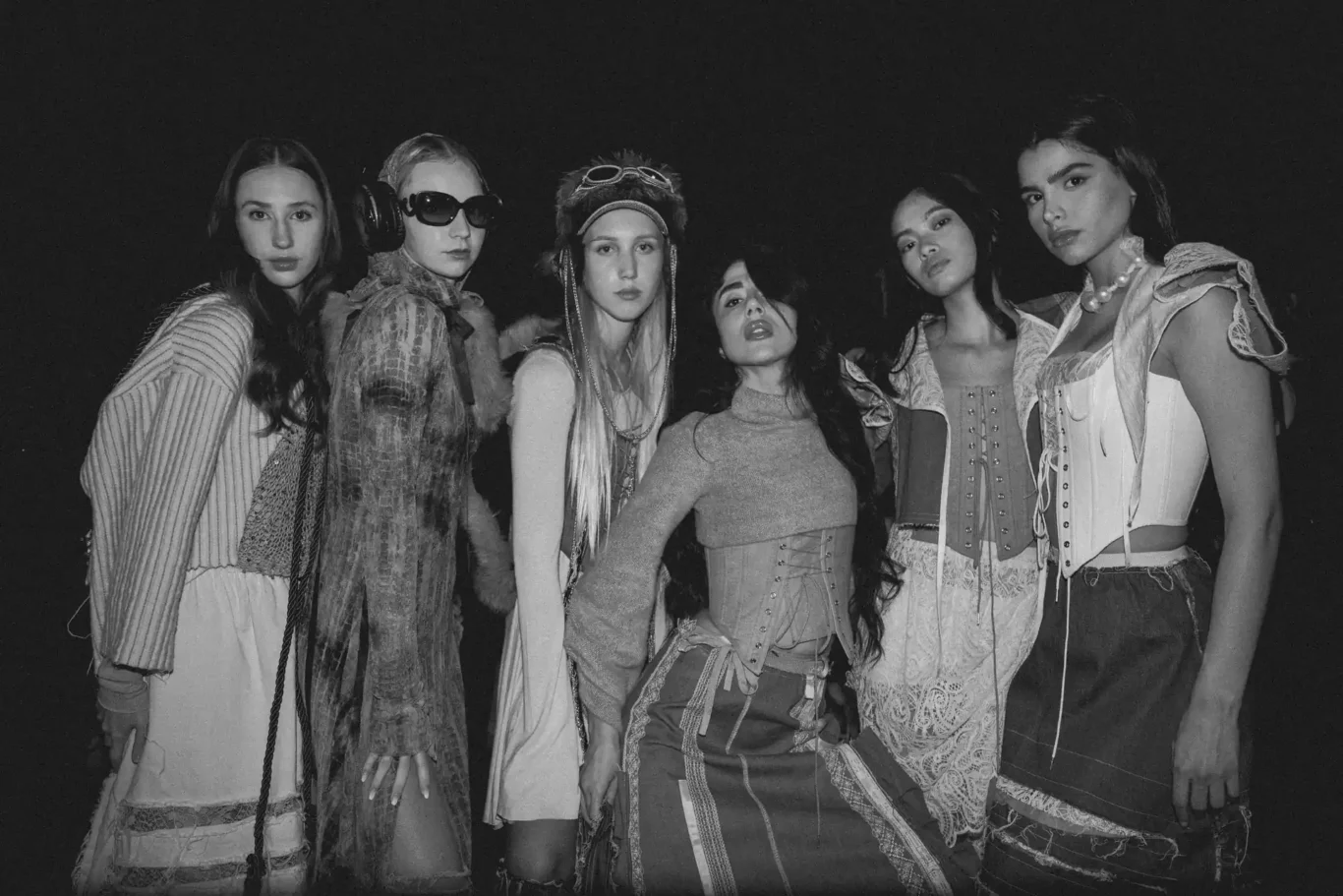 Six women in vintage-inspired outfits pose together for a monochrome fashion shoot at night, serious expressions at the camera.