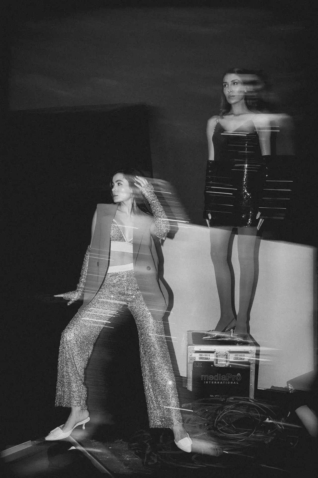 Two female models pose on a backstage fashion set, one in a glittery jumpsuit seated and the other in a black sequined dress standing, with motion blur suggesting movement and lighting equipment nearby.