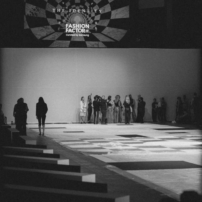 Black-and-white fashion runway scene with a group of people gathered at the far end; a large screen displays 'THE IDENTITY' and 'FASHION FACTOR'.