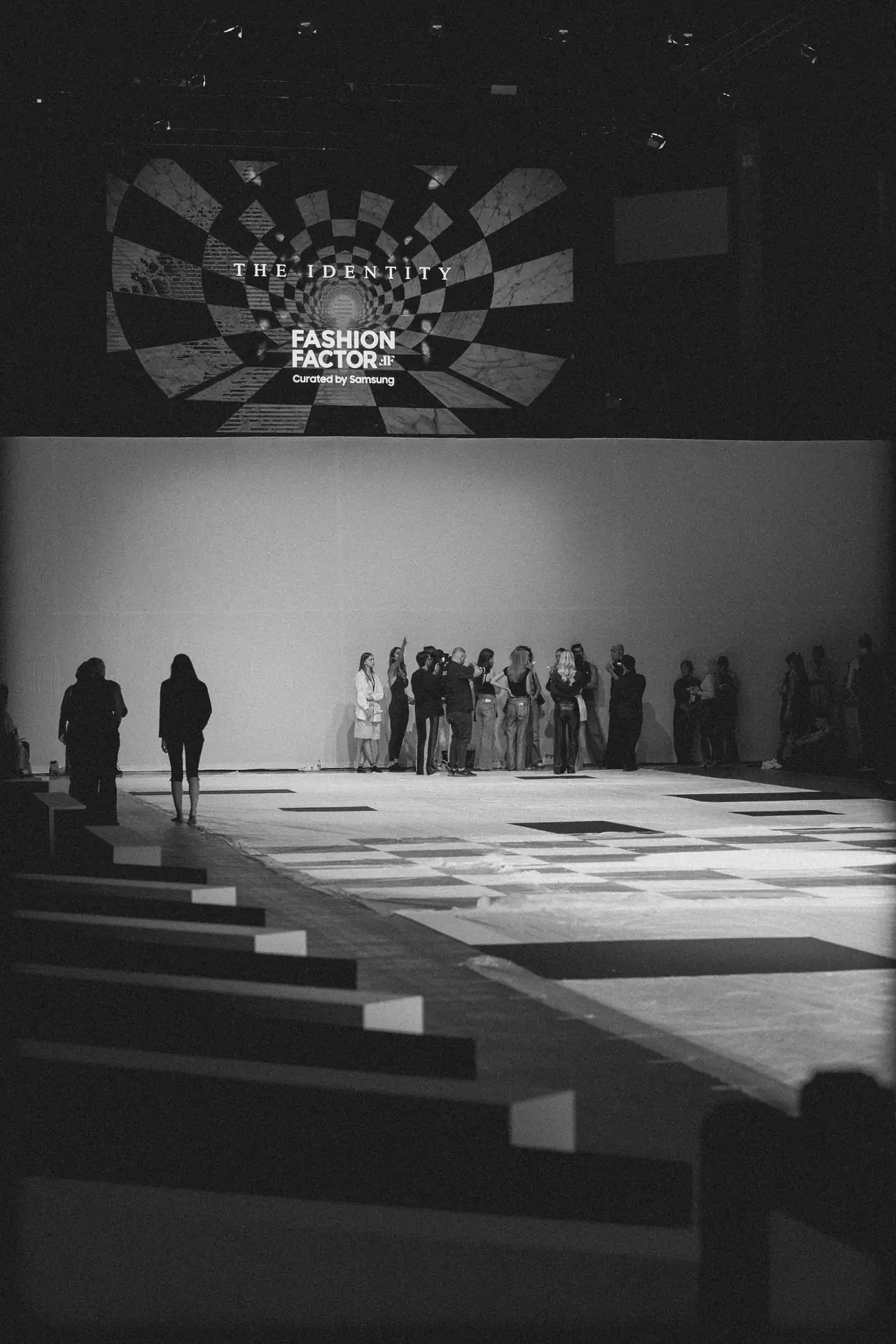 Black-and-white fashion runway scene with a group of people gathered at the far end; a large screen displays 'THE IDENTITY' and 'FASHION FACTOR'.
