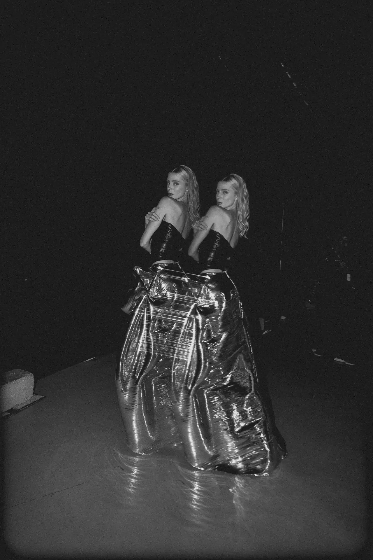 Two women with wavy blonde hair pose back-to-back in black strapless outfits on a large, shiny metallic gown outdoors at night.