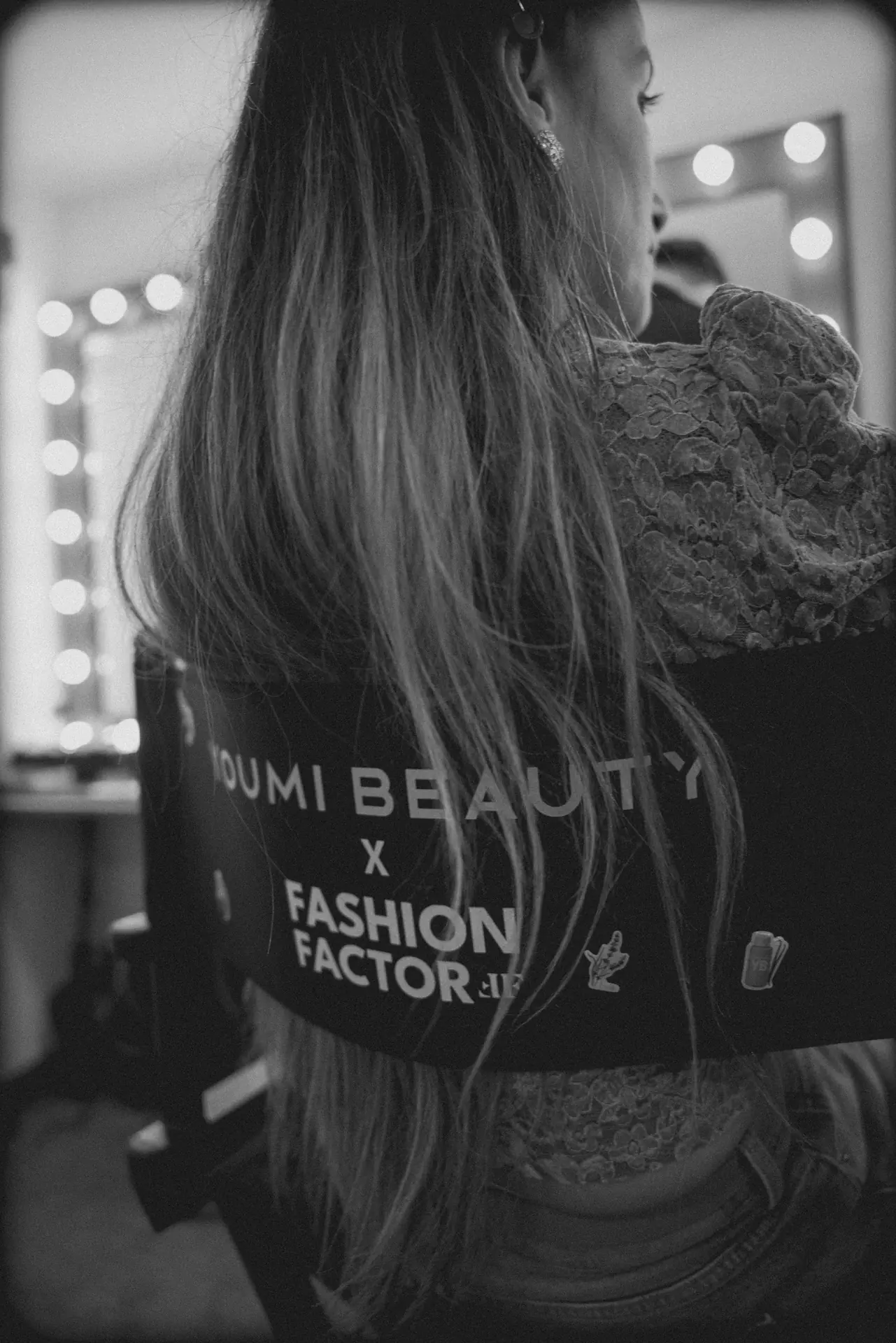 Back view of a person with long hair, wearing a lace top, in a dressing room with round vanity lights in the background