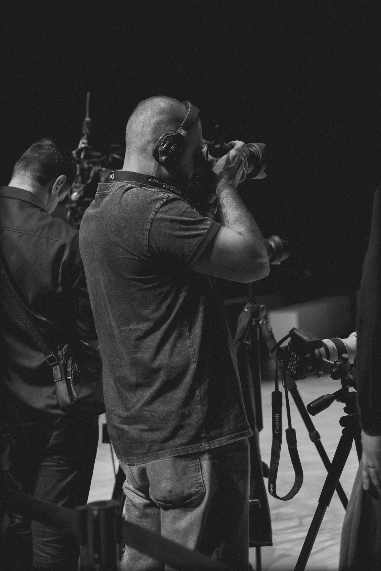 Photographer in a studio aiming a long lens at a subject, wearing headphones, with other gear and a second person in the background (black-and-white image).
