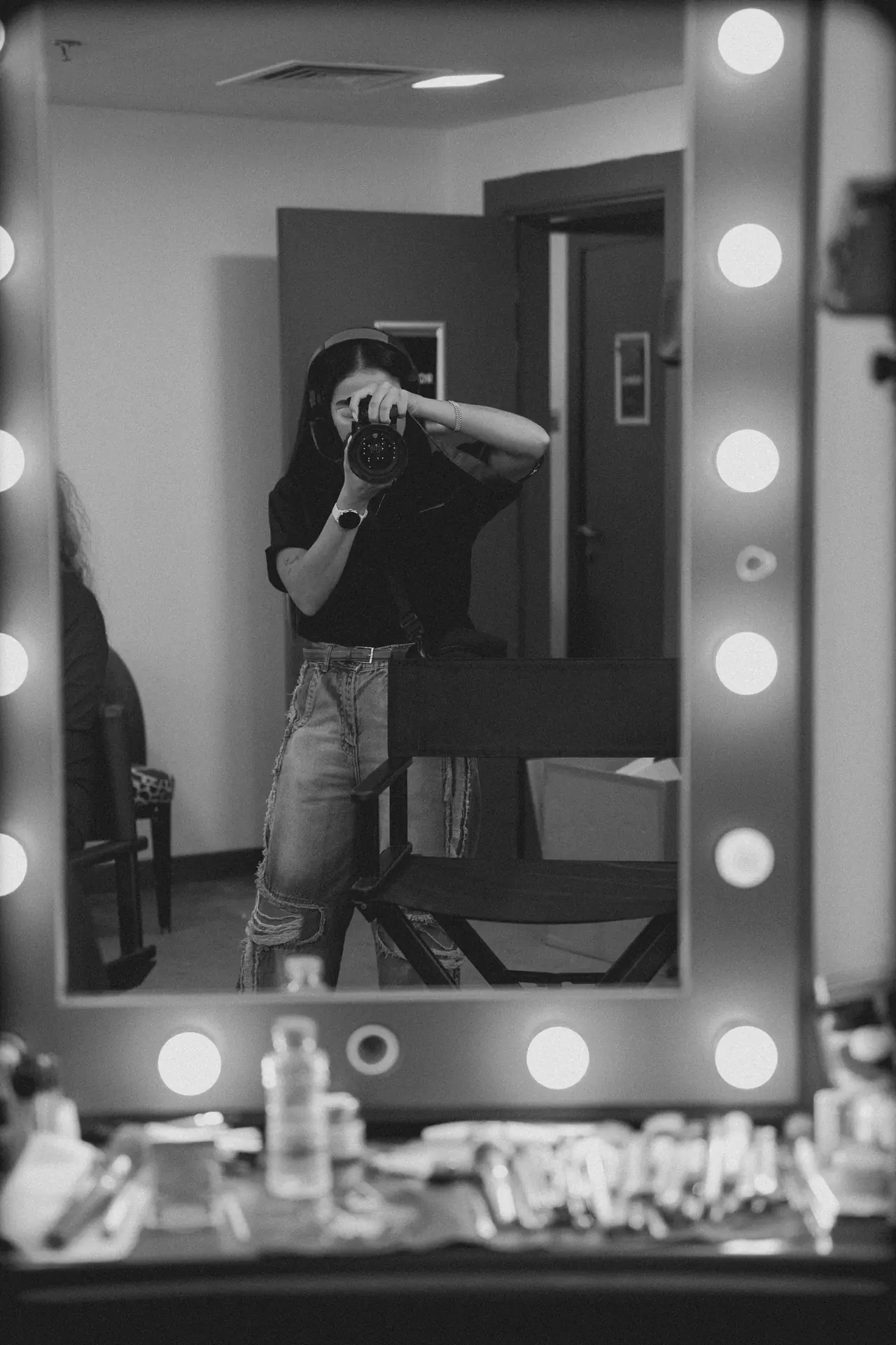 Person in a black shirt and jeans takes a self-portrait in a vanity mirror, holding a camera to their face.