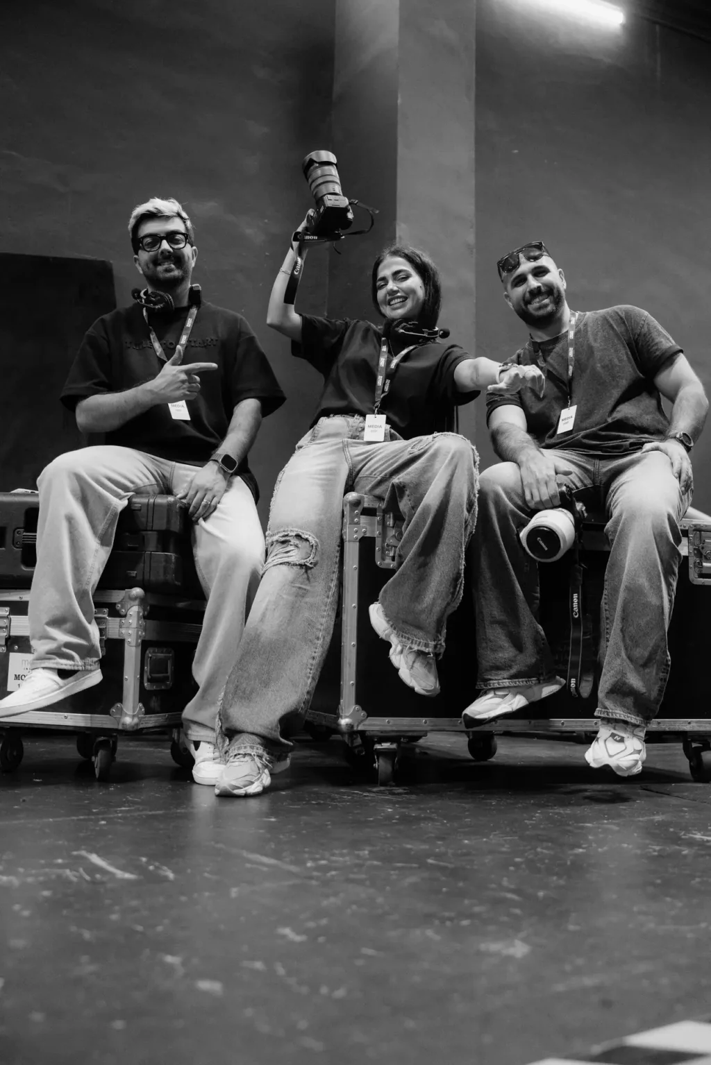 Three media crew members sit on equipment cases, smiling; the person in the center holds a large lens overhead, while the others have cameras and lanyards in a studio setting.