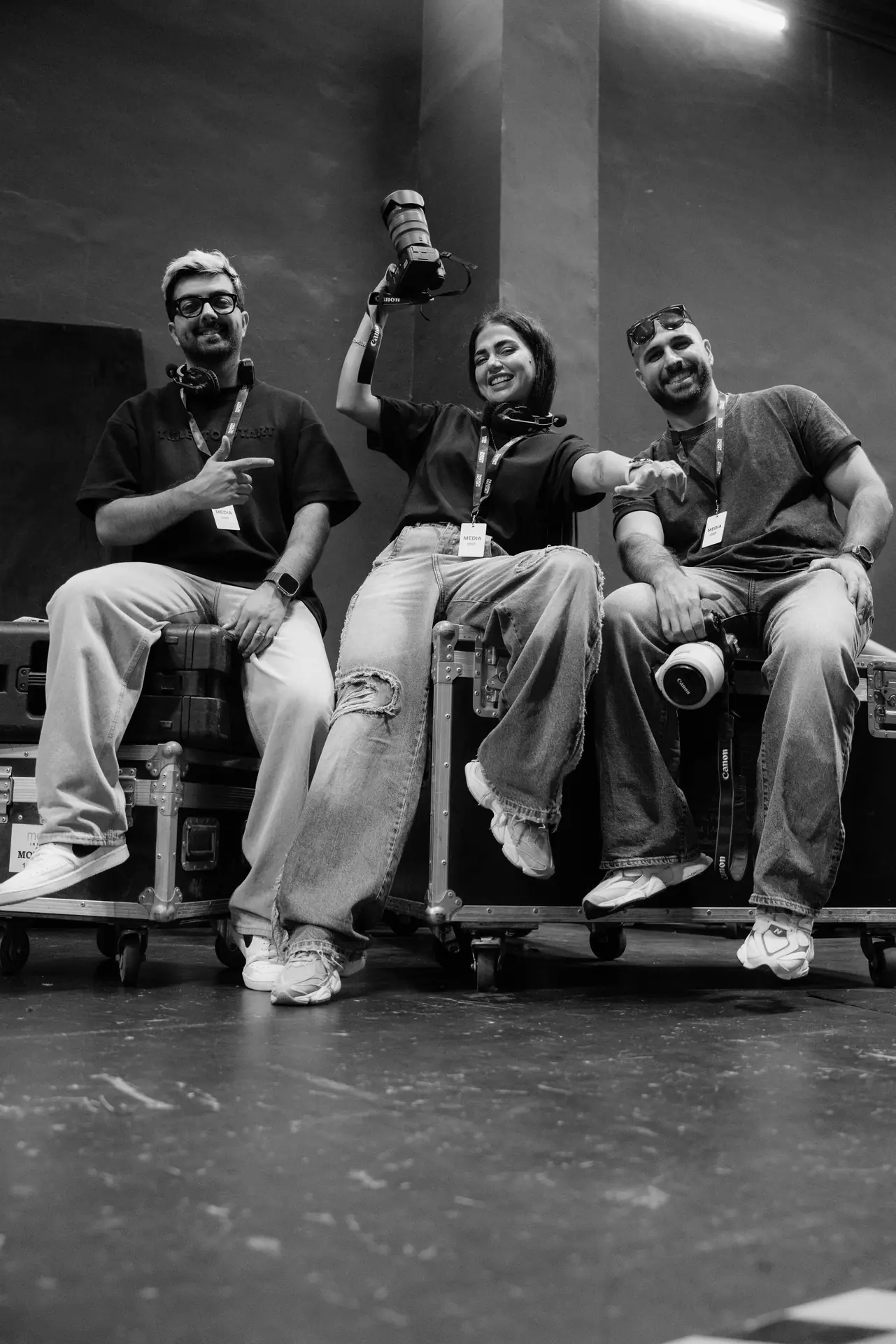 Three media crew members sit on equipment cases, smiling; the person in the center holds a large lens overhead, while the others have cameras and lanyards in a studio setting.