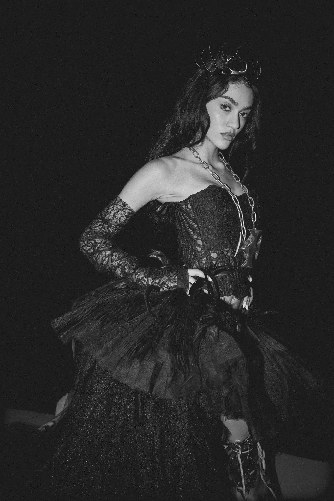 Black-and-white fashion portrait of a woman in a lace dress with a chunky chain necklace and crown against a dark background.