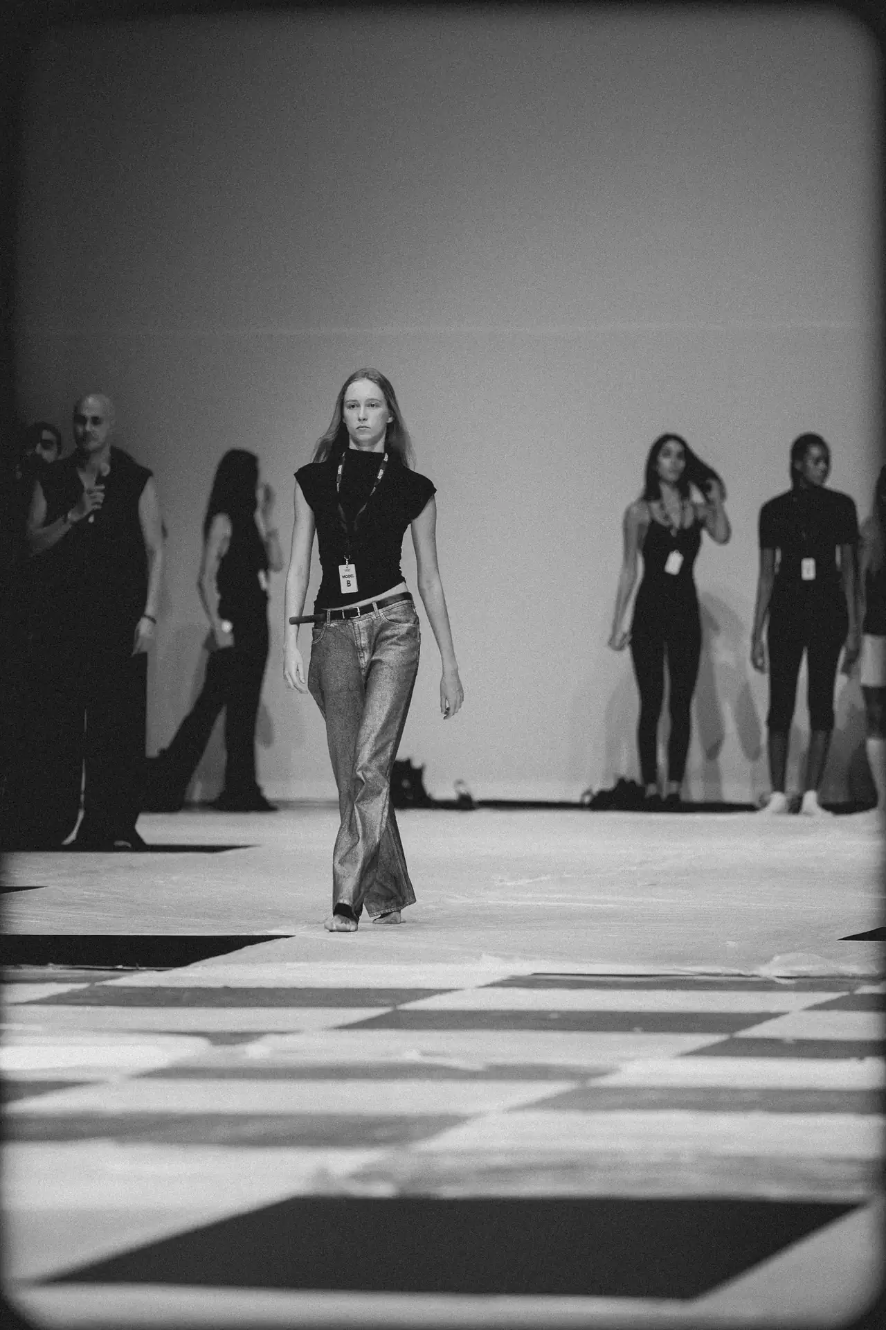 Model walks down a fashion runway in a sleeveless black top and jeans, with backstage figures in the background.