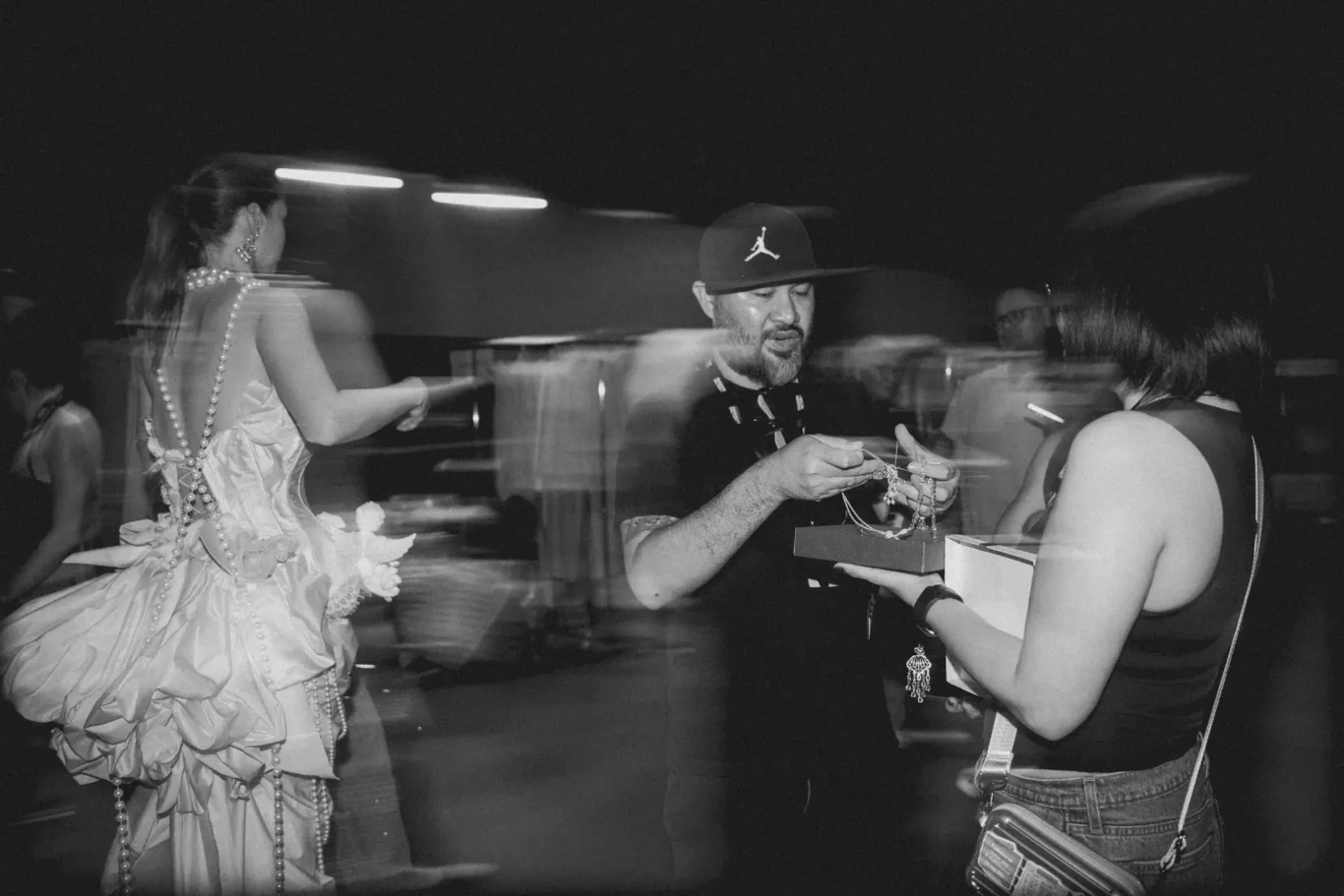 Man in a baseball cap hands jewelry to a woman in a voluminous ball gown at a dark, festive event.