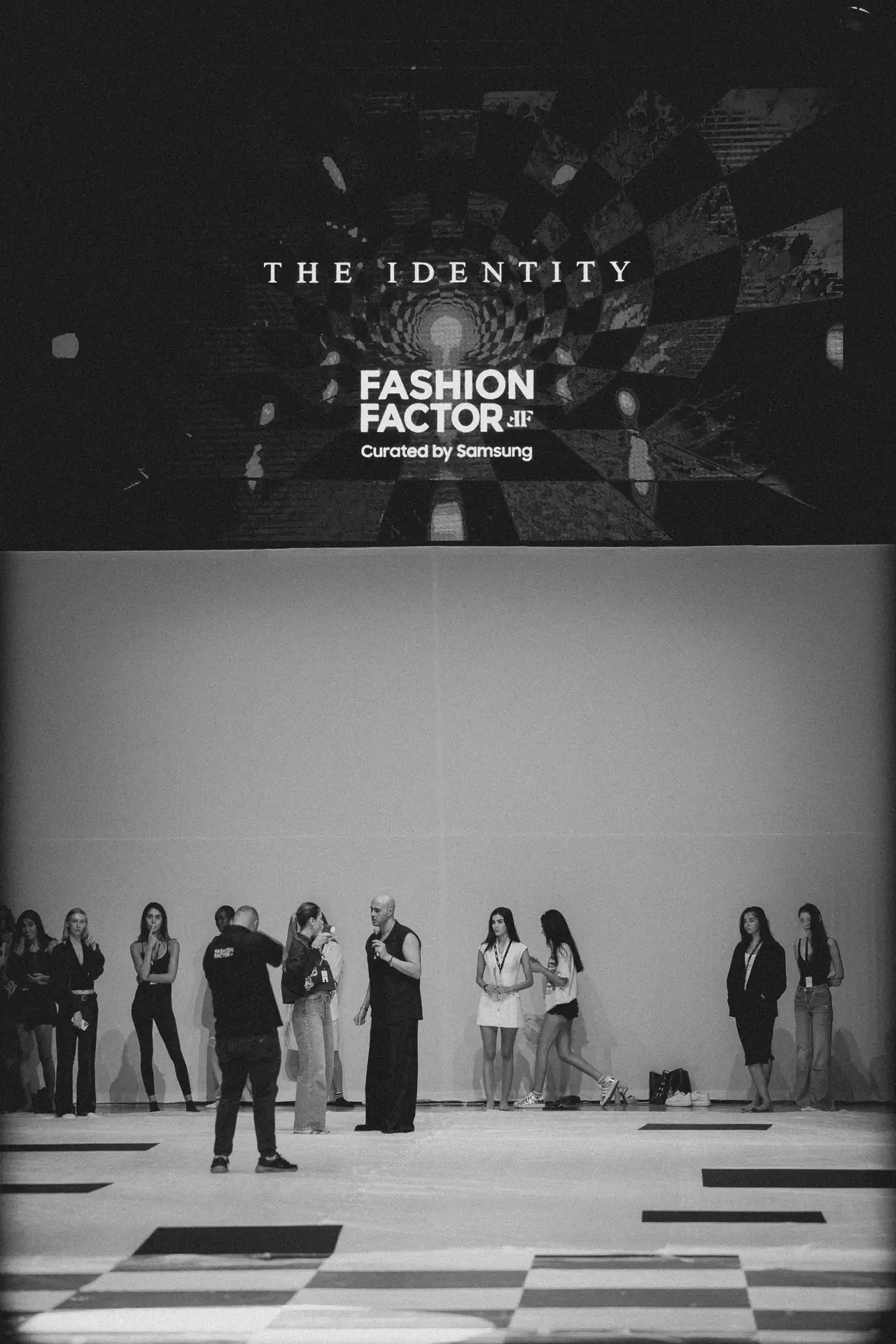 Models line up on a runway during a fashion event; large screen behind reads 'THE IDENTITY' and 'FASHION FACTOR' with 'Curated by Samsung' text.