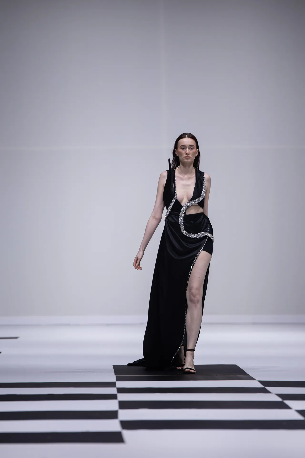 Model walks a black evening gown with silver beaded trim along a high-contrast black-and-white runway.