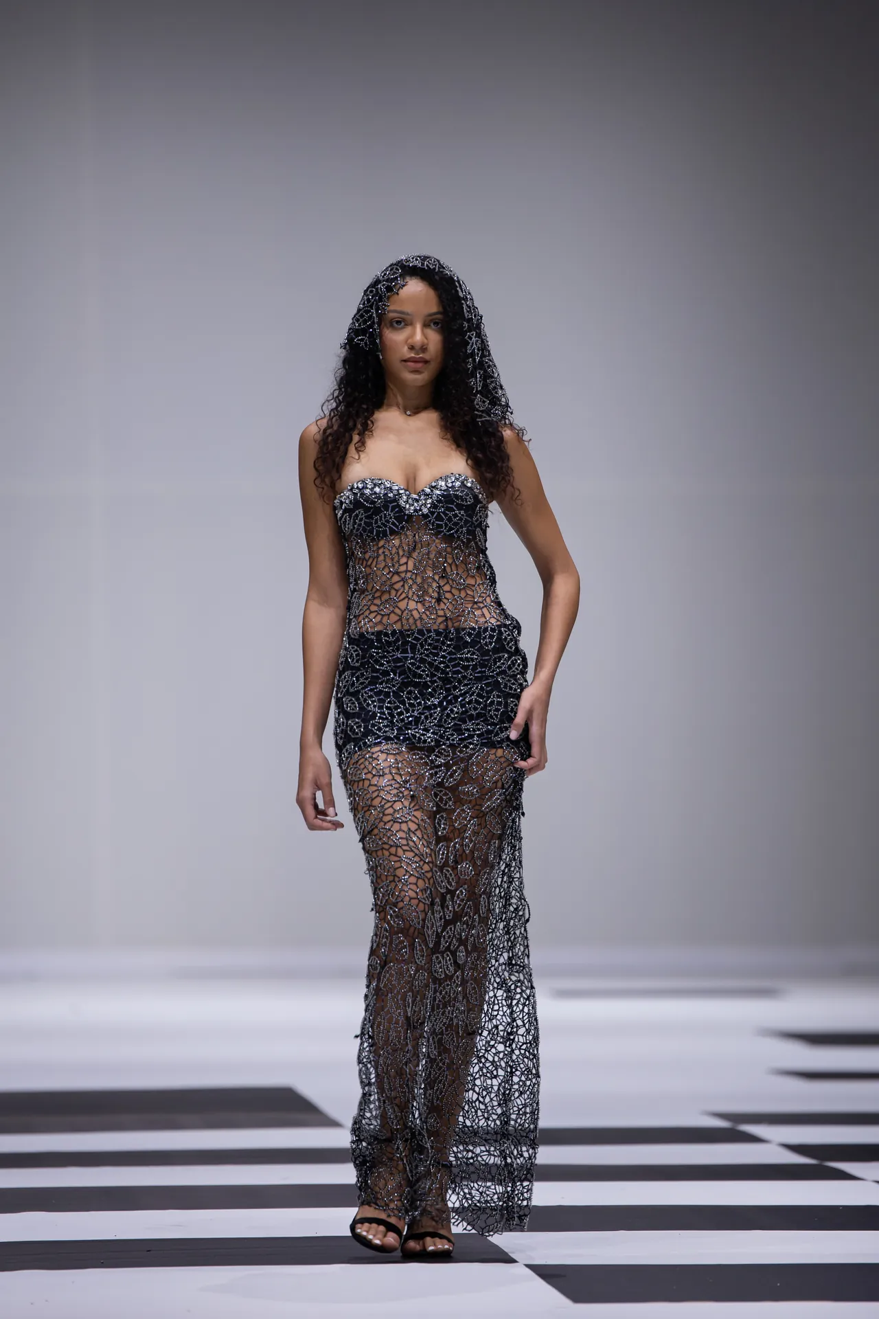 Female model on a runway wearing a black sheer beaded gown with a geometric mesh pattern, walking toward the camera.