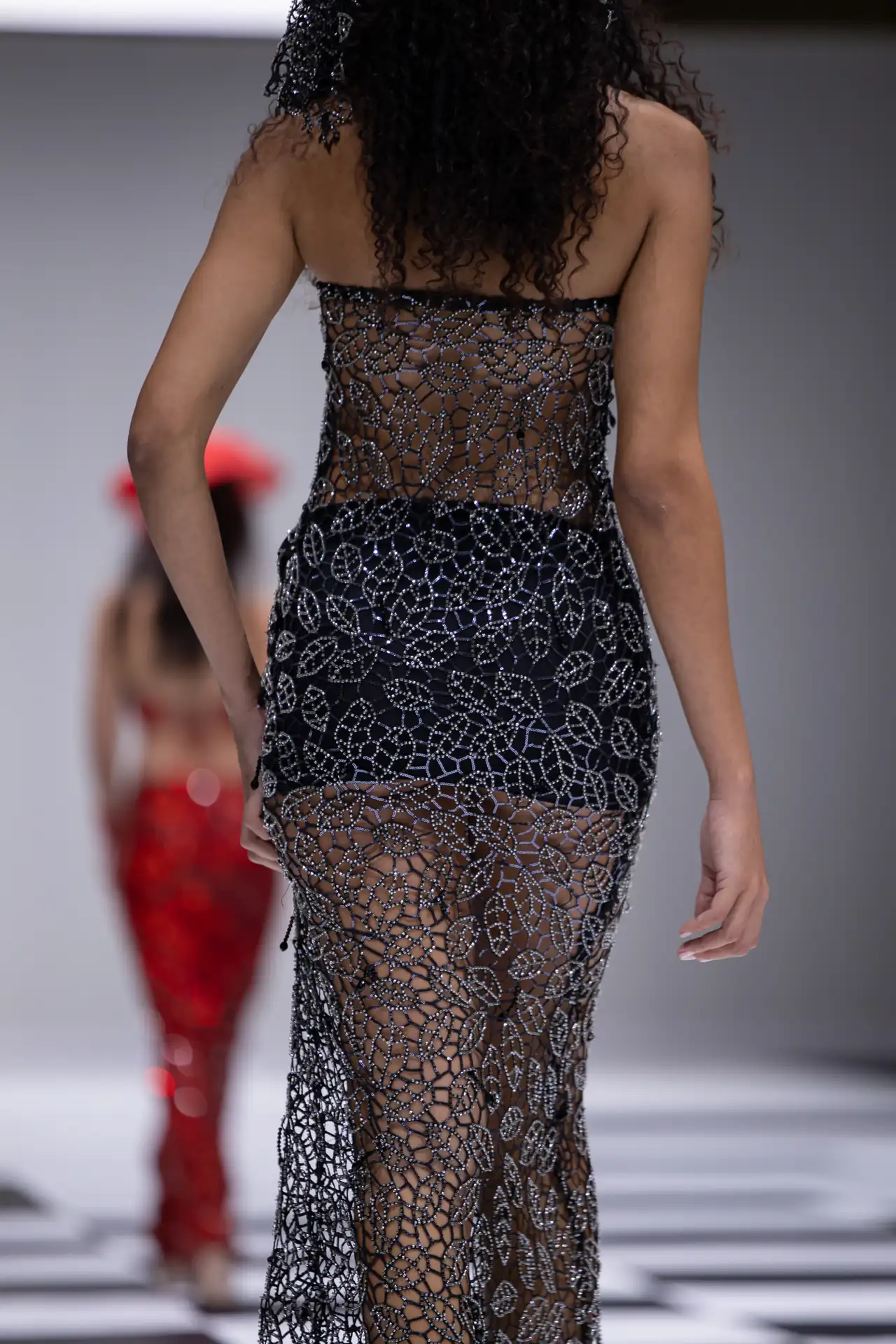 Back view of a model wearing a sheer black gown with silver beaded lattice overlay on the runway.