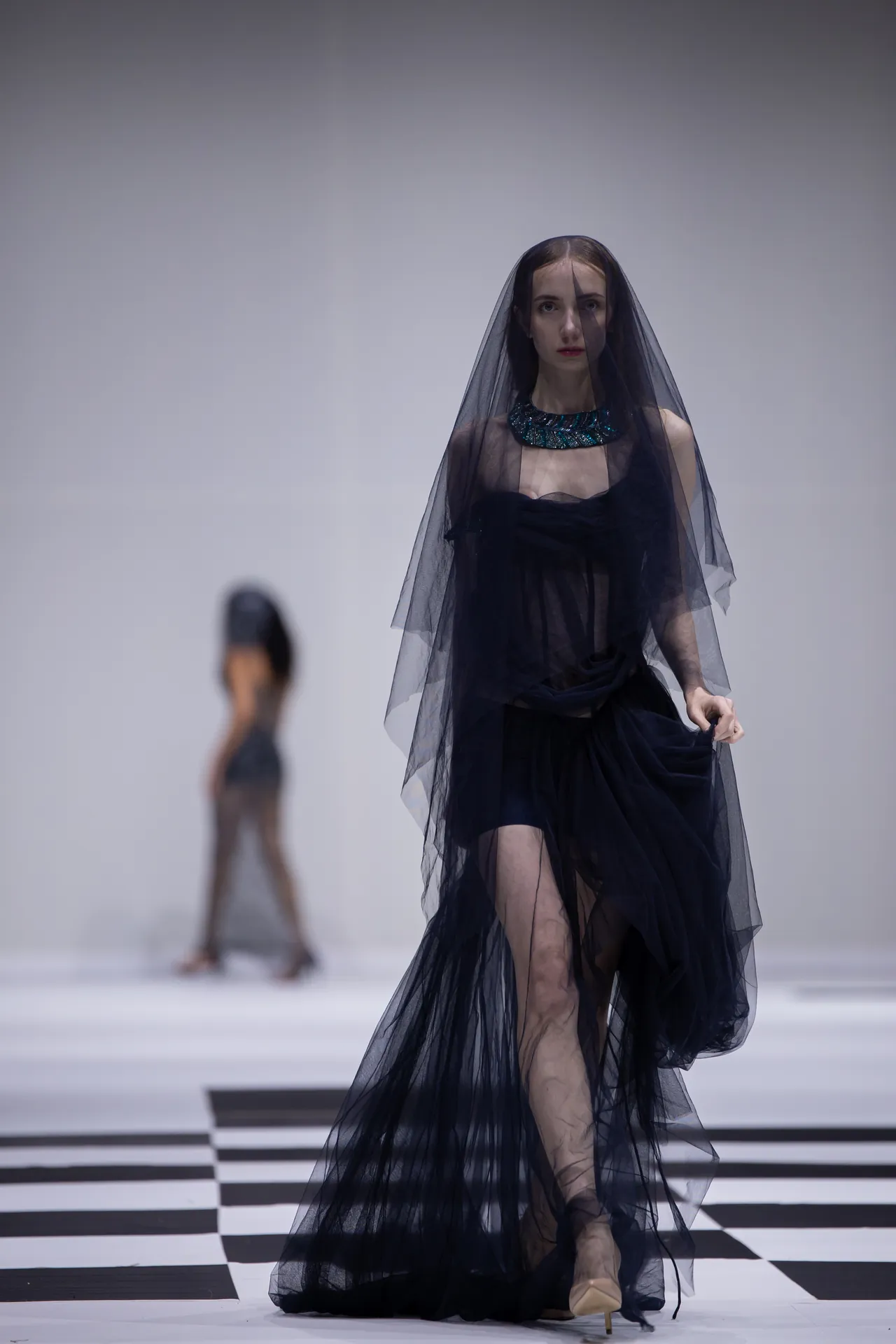 Model on a runway wearing a black sheer gown with a teal beaded collar and veil, walking over a black-and-white checkered floor.