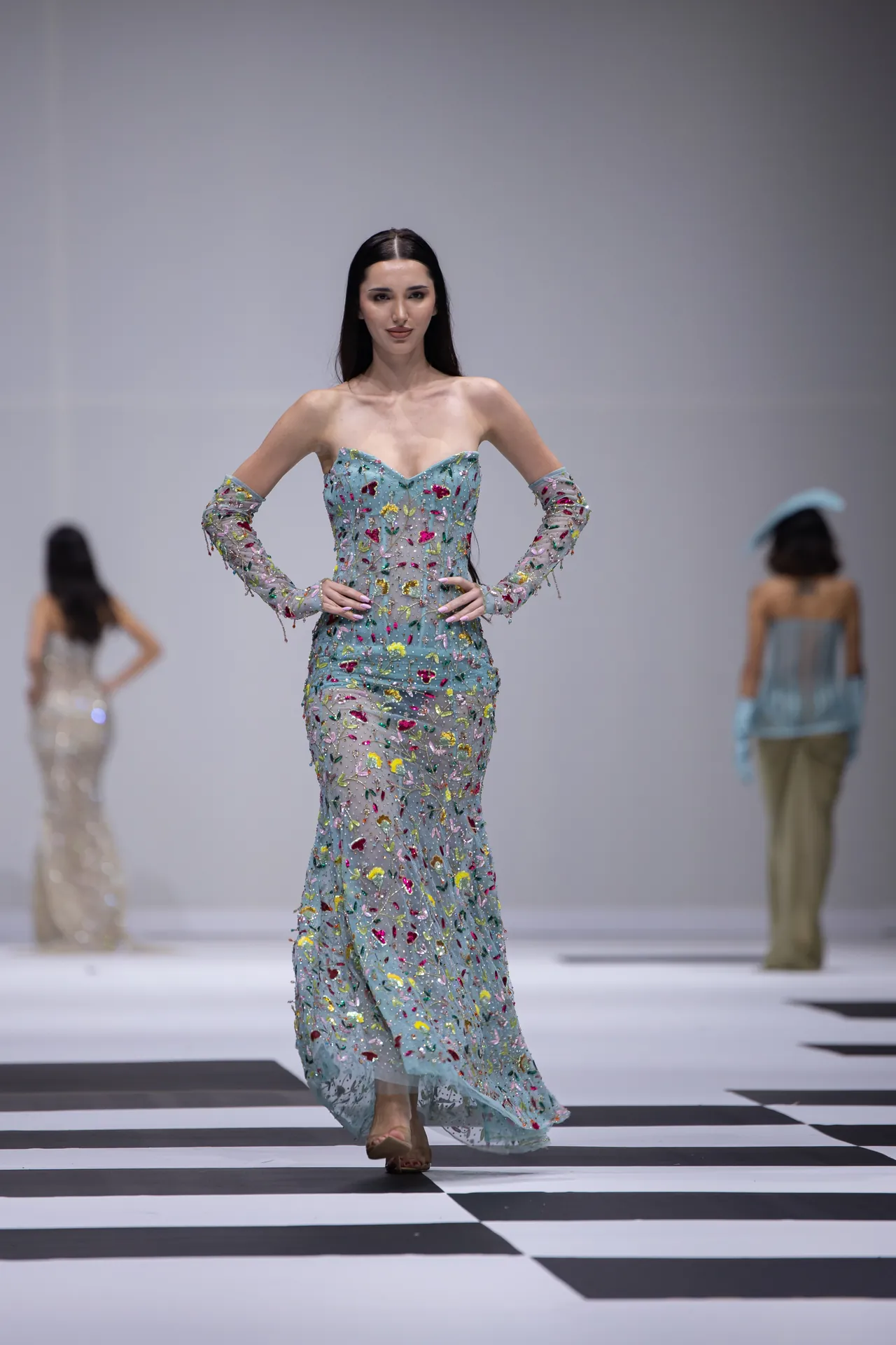 Model walks a runway wearing a light blue off-shoulder gown with colorful floral embroidery and sheer long sleeves.