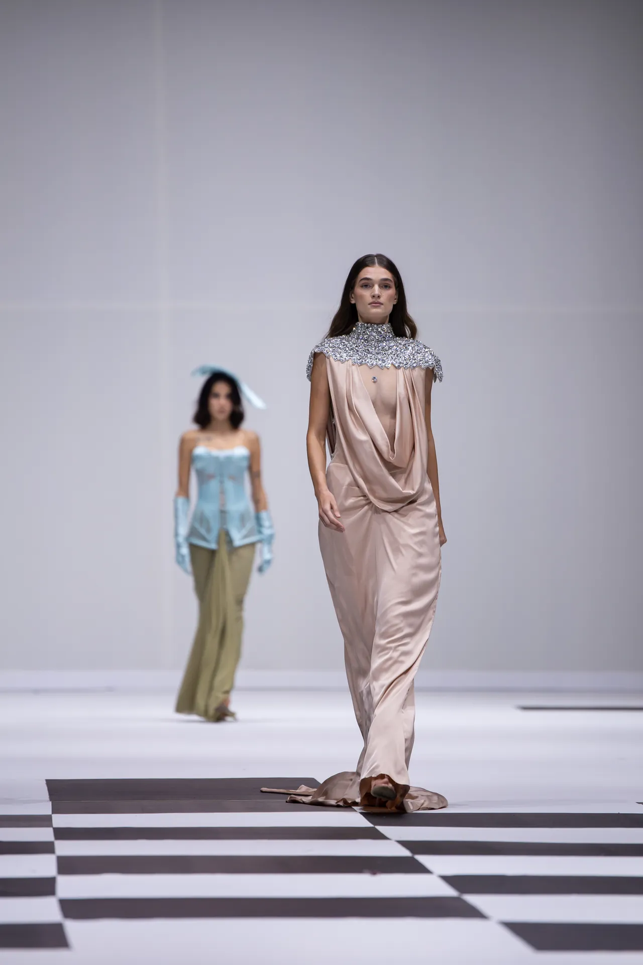 Model on a runway wearing a pale pink satin gown with a jeweled collar; another model in a blue corset and khaki pants is blurred in the background on a black-and-white checkered floor.