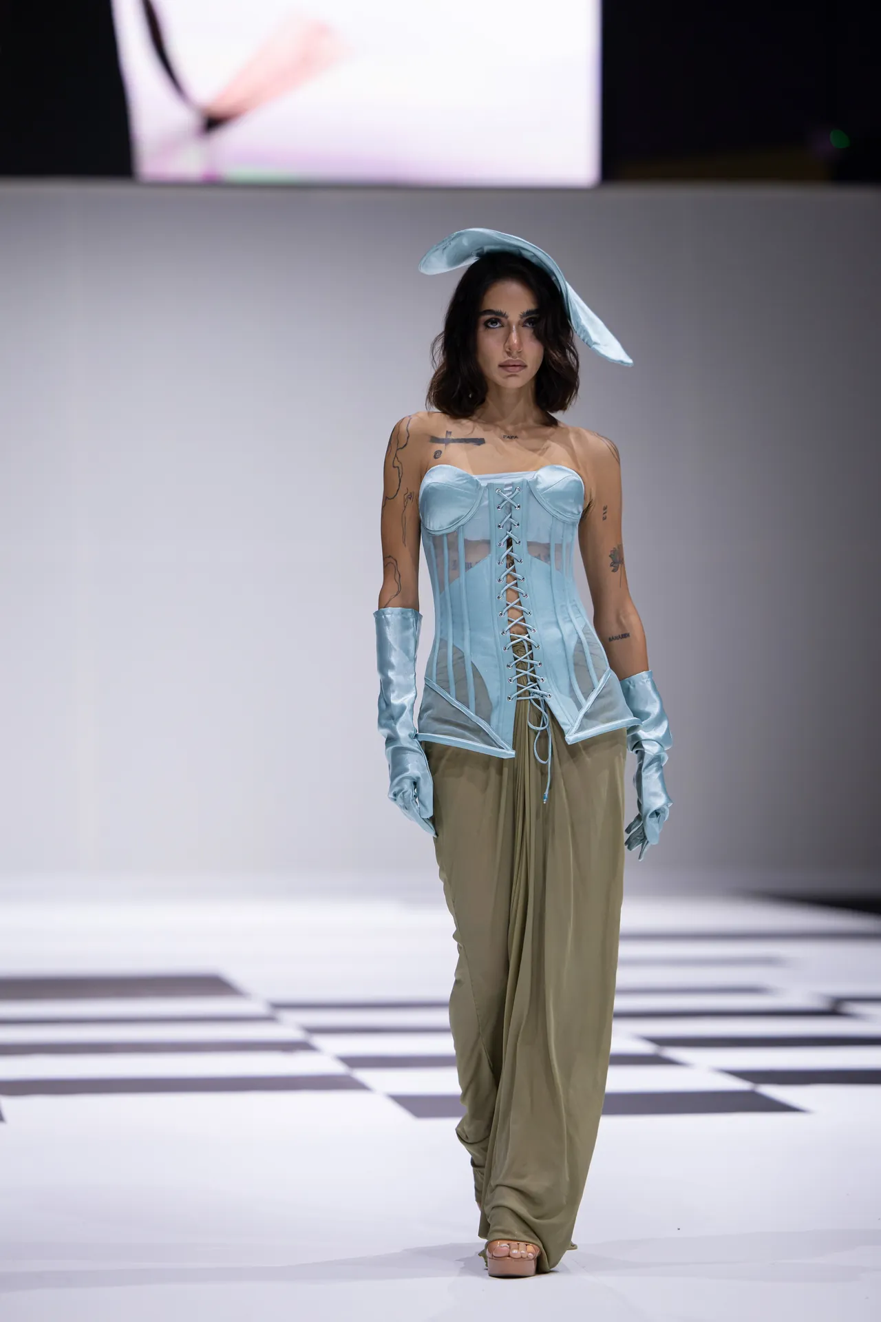 Model walks the runway in a pale blue corset with lace-up front and sheer panels, paired with satin gloves and a matching hat.