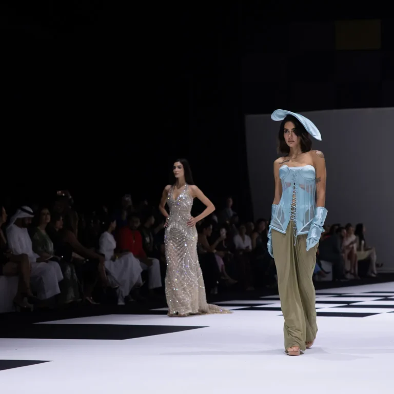Model walks a fashion runway wearing a light blue corseted top with sheer panels and beige wide-leg pants; large blue hat on her head; bold black-and-white checkered floor and dark audience background.