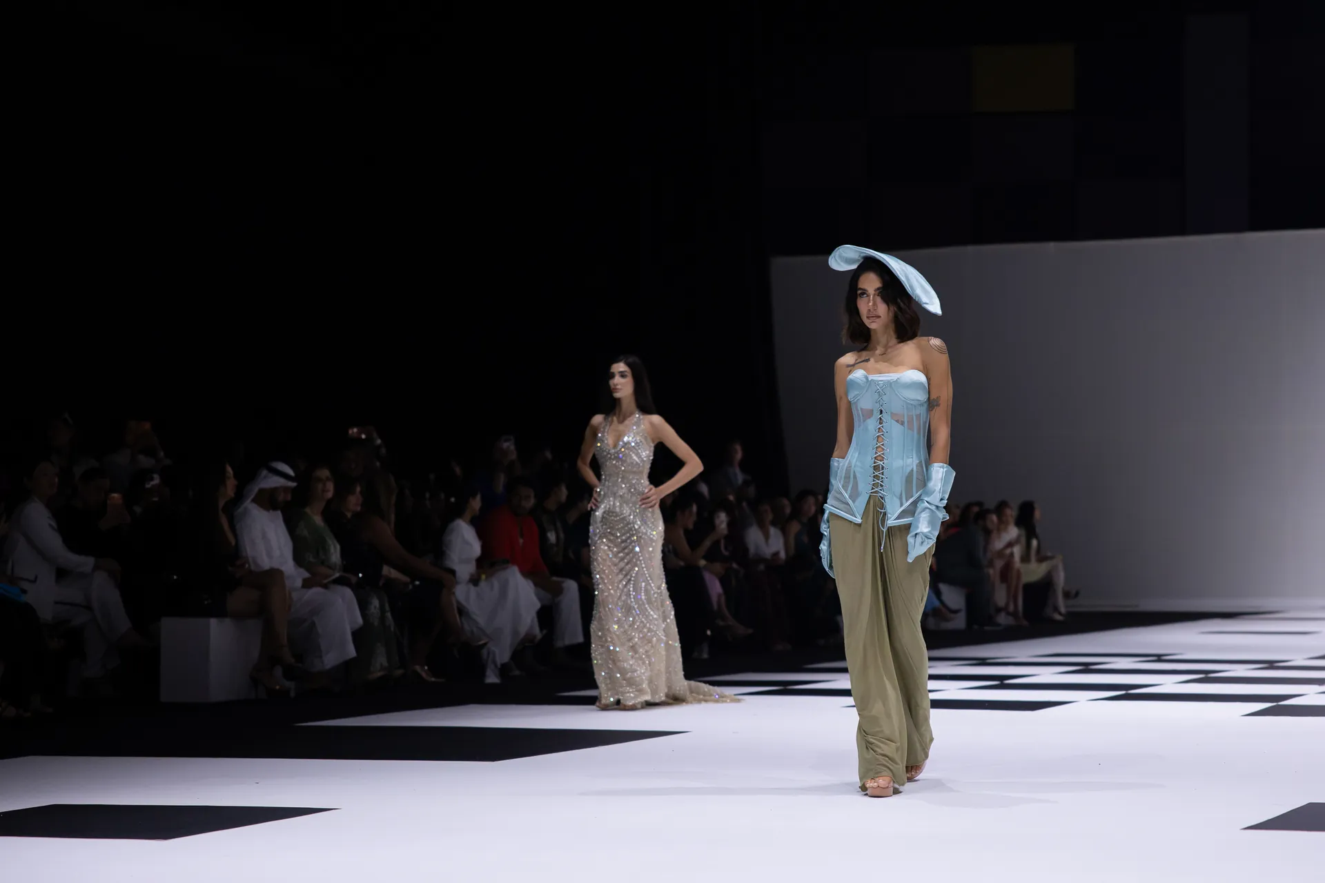 Model walks a fashion runway wearing a light blue corseted top with sheer panels and beige wide-leg pants; large blue hat on her head; bold black-and-white checkered floor and dark audience background.
