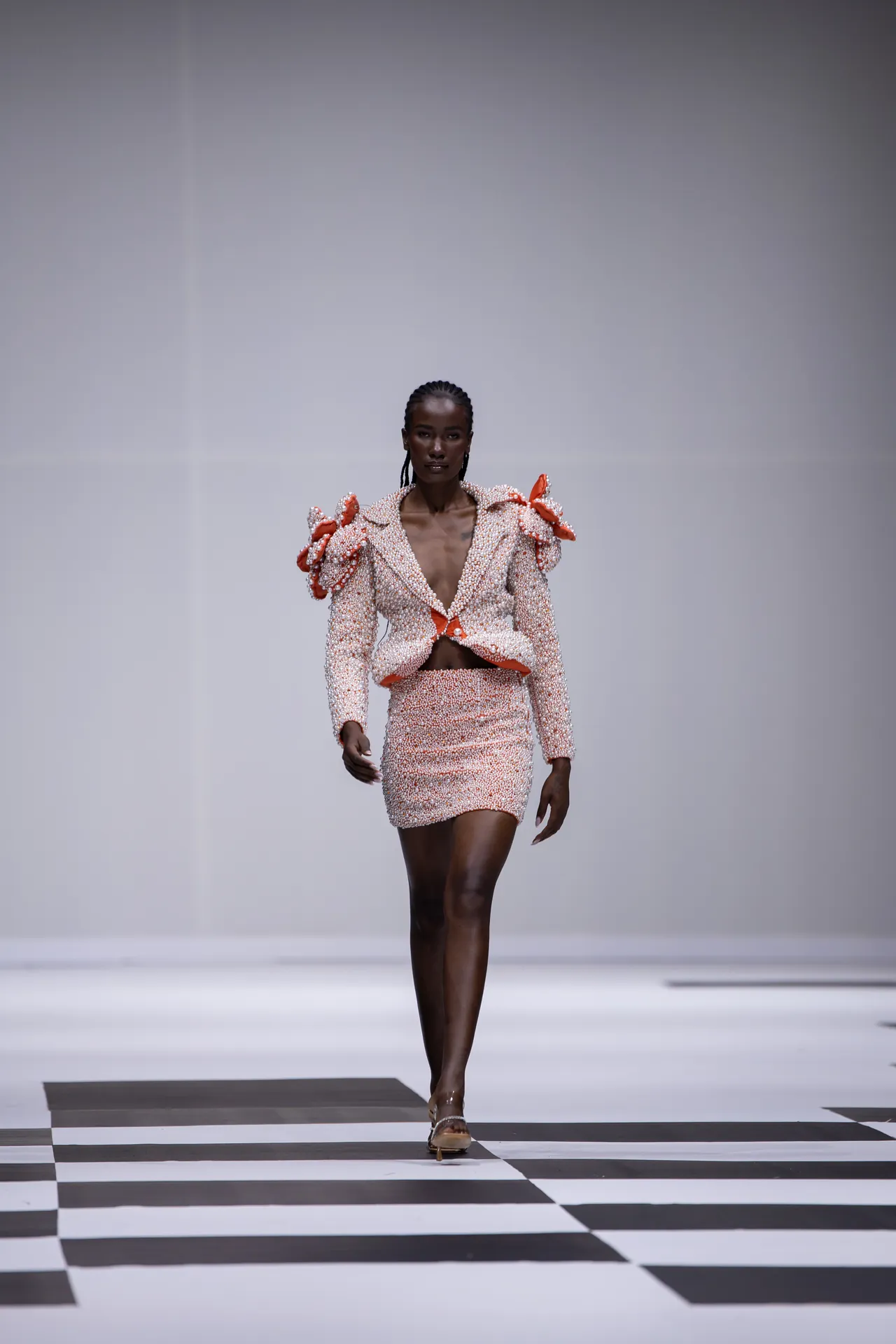 Model walks a fashion show wearing a pearl-beaded pink suit with orange ribbon accents and puff shoulders on a black-and-white checkered runway.