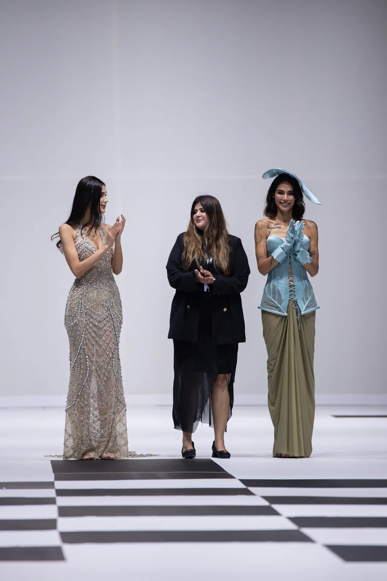 Three models stand on a black-and-white checkered runway; left in a sparkling silver gown, center in a black blazer, right in a light blue corset and gloves.