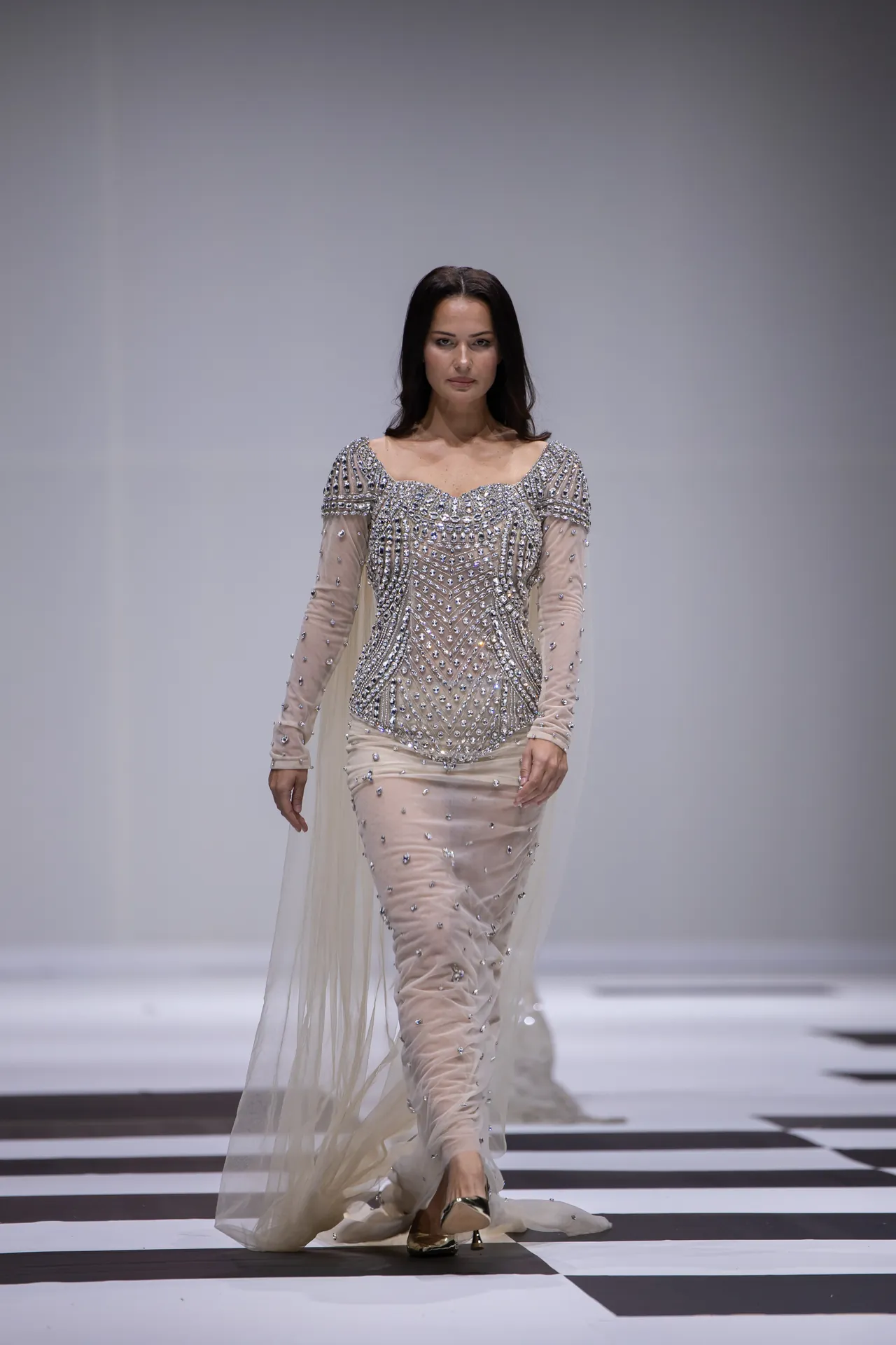 Model walking a runway in a form-fitting champagne gown with heavy silver beading and sheer sleeves or cape, on a light backdrop.