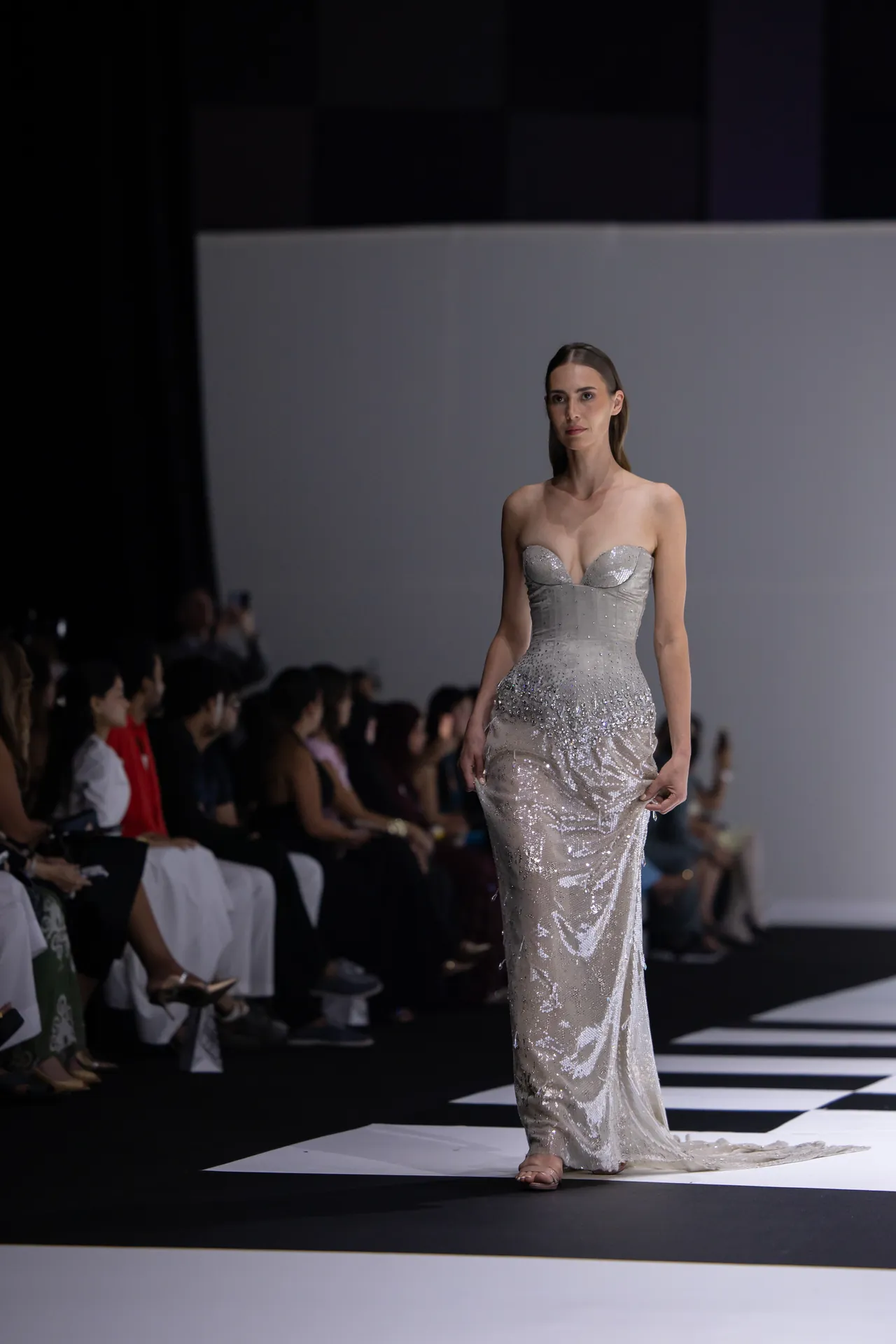 Model walks the runway in a silver sequined strapless gown as the audience watches in the background.