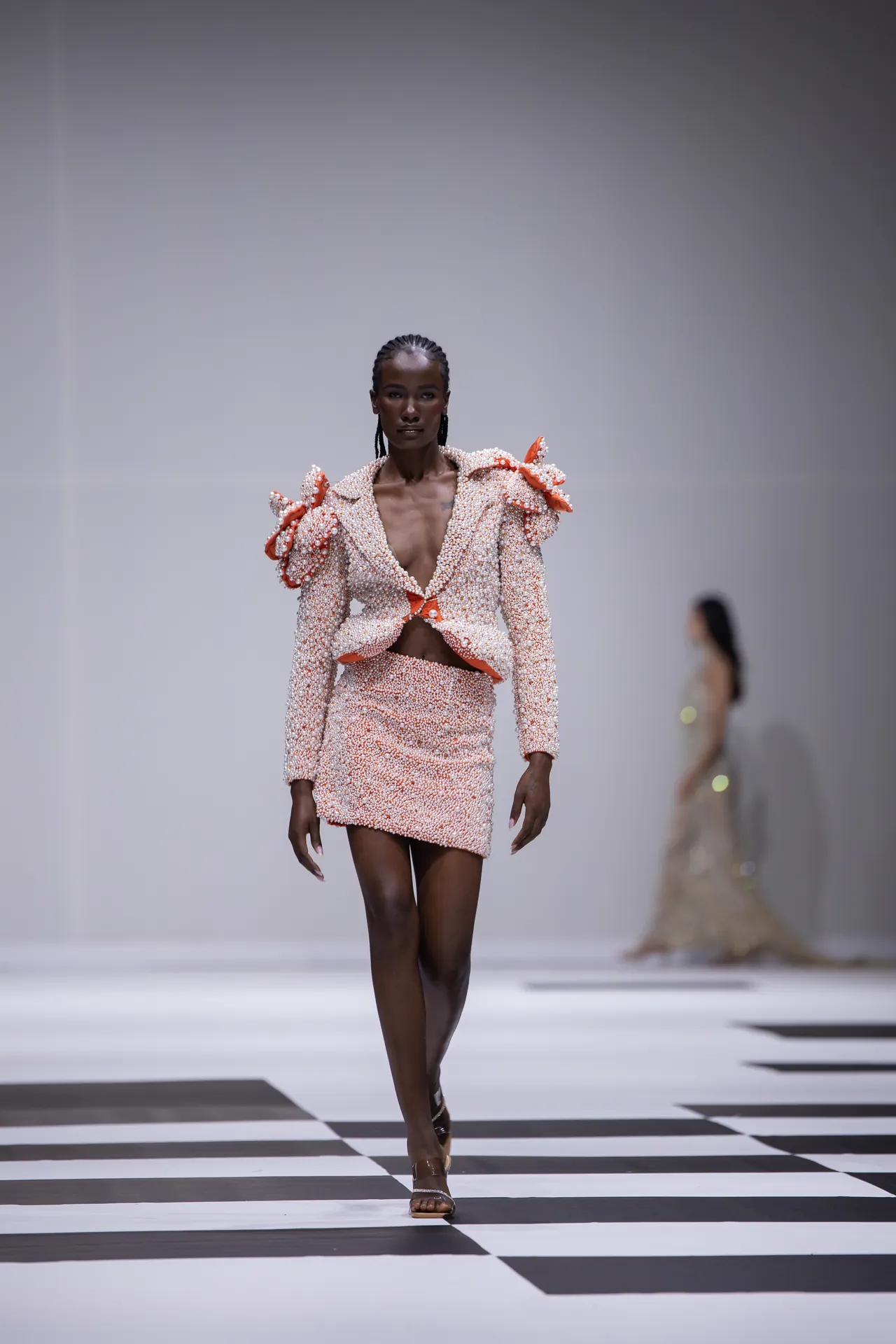 Model walks the runway in a pearl-studded pink suit with oversized shoulders on a black-and-white checkered catwalk, with another model blurred in the background.