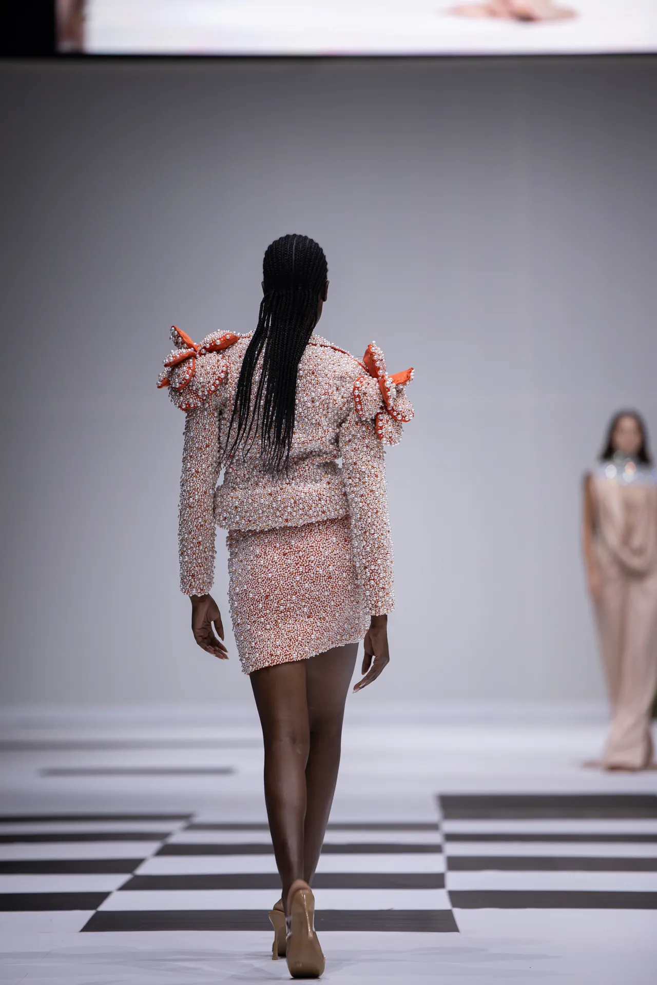 Back view of a model walking the runway in a pink bead-covered suit with fluffy orange shoulder details.