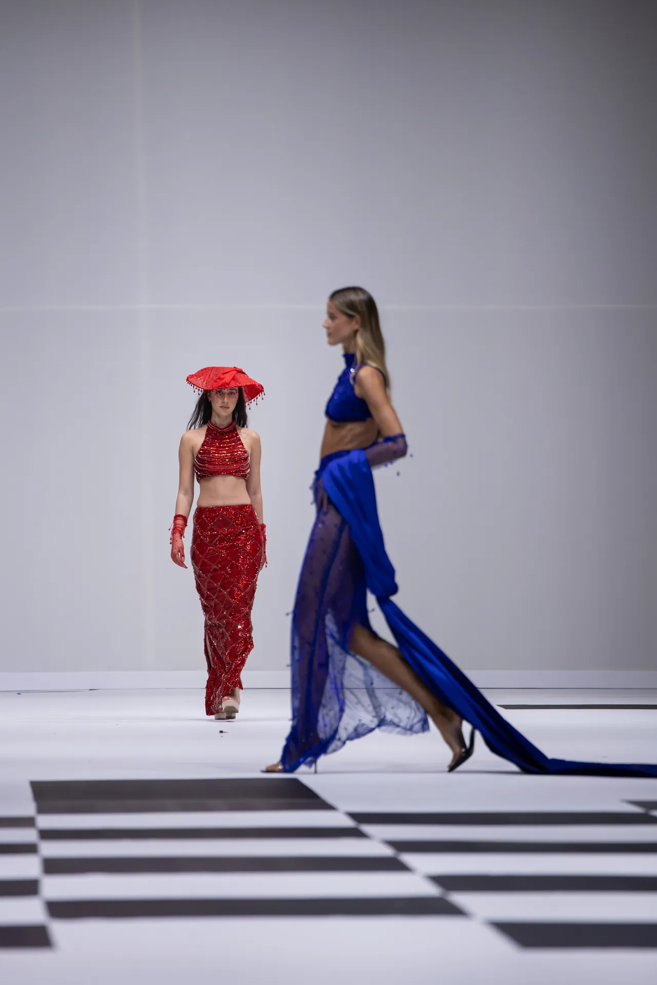 Two models walk a fashion runway: one in a red sequined two-piece with a matching red hat, the other in a flowing blue gown on a white catwalk with black-and-white foreground stripes.