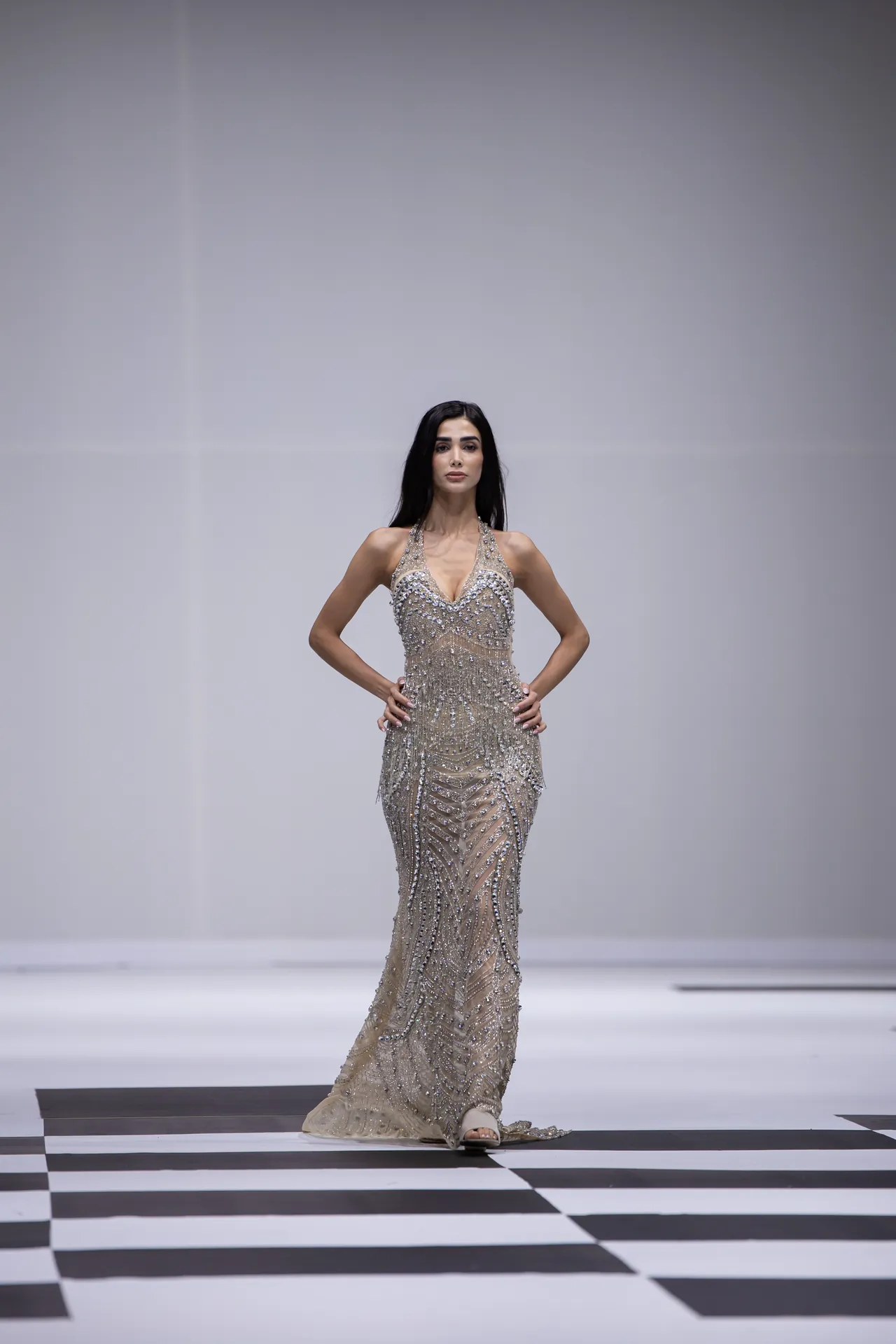 Female model walks a glamorous sequined evening gown on a black-and-white checker runway, hands on hips.