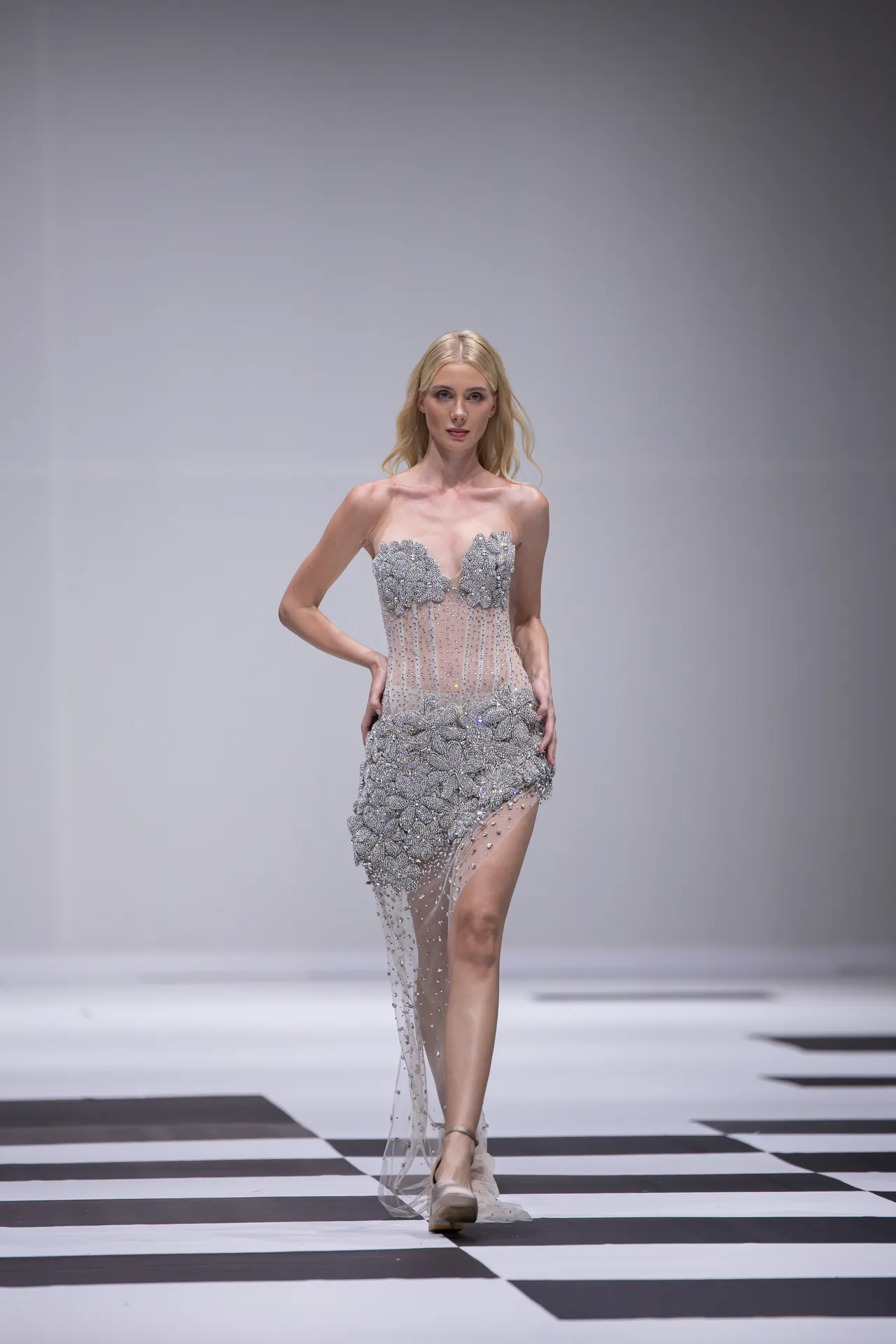 Female model walks a silver, beaded gown with floral appliqués on a black-and-white checkered runway.