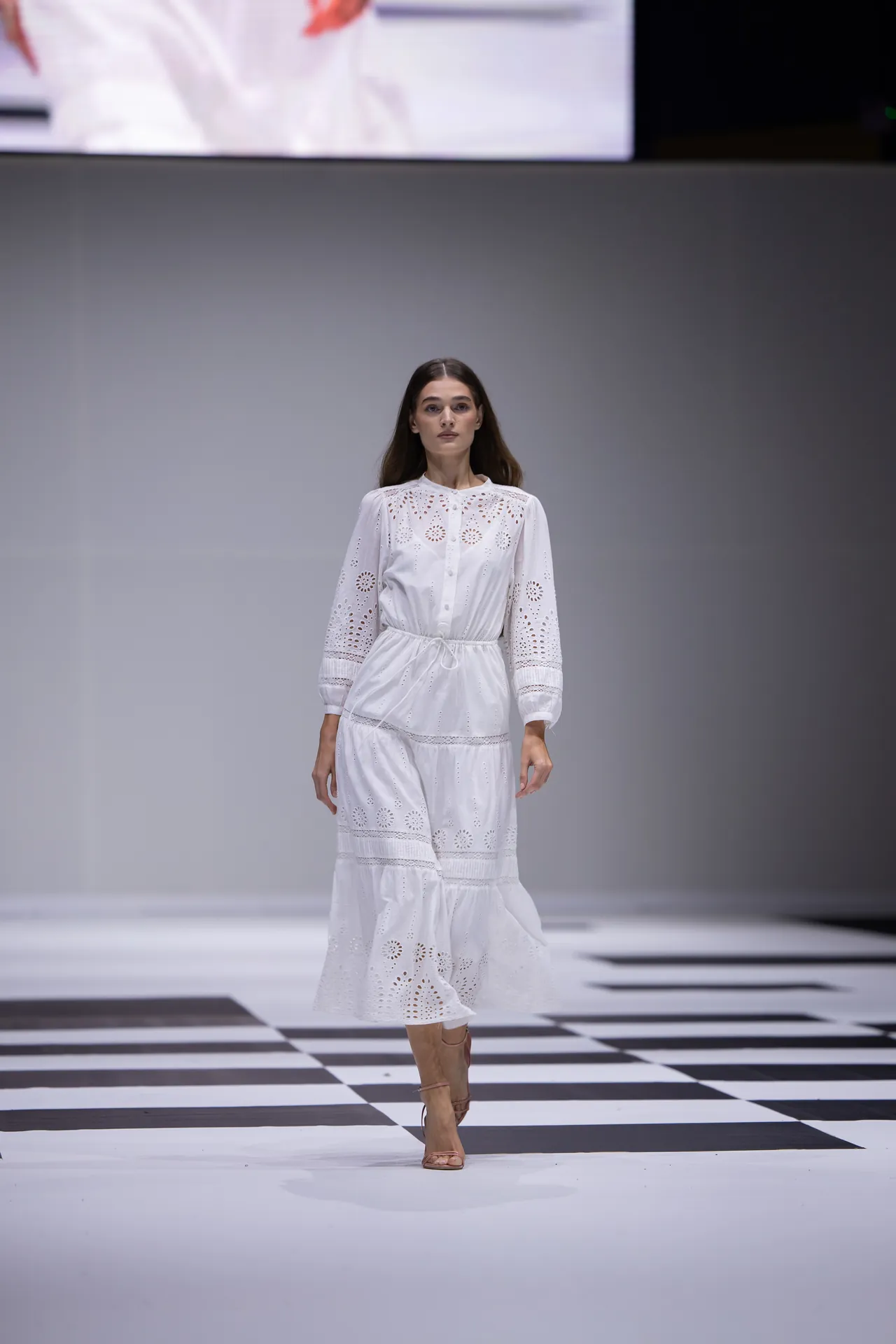 Female model walking down a checkerboard runway in a white eyelet lace dress.