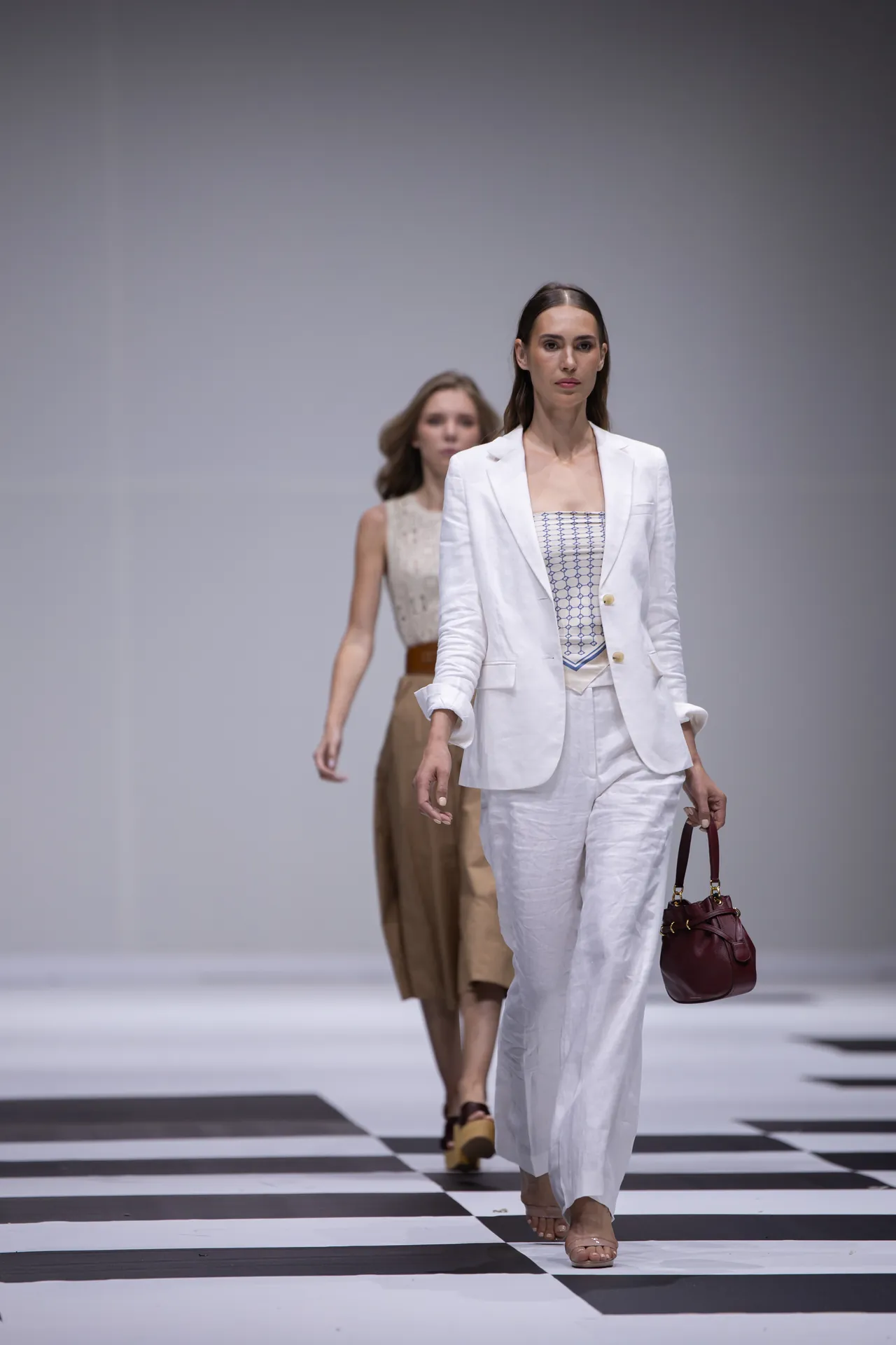 Model in a white suit walks a fashion runway, holding a burgundy handbag, with a black-and-white checkered floor.”] ,