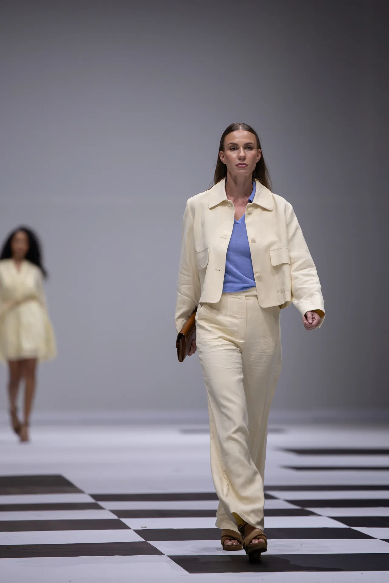 Model walks the runway wearing a cream suit with a light blue top, carrying a brown clutch on a black-and-white checkered floor.