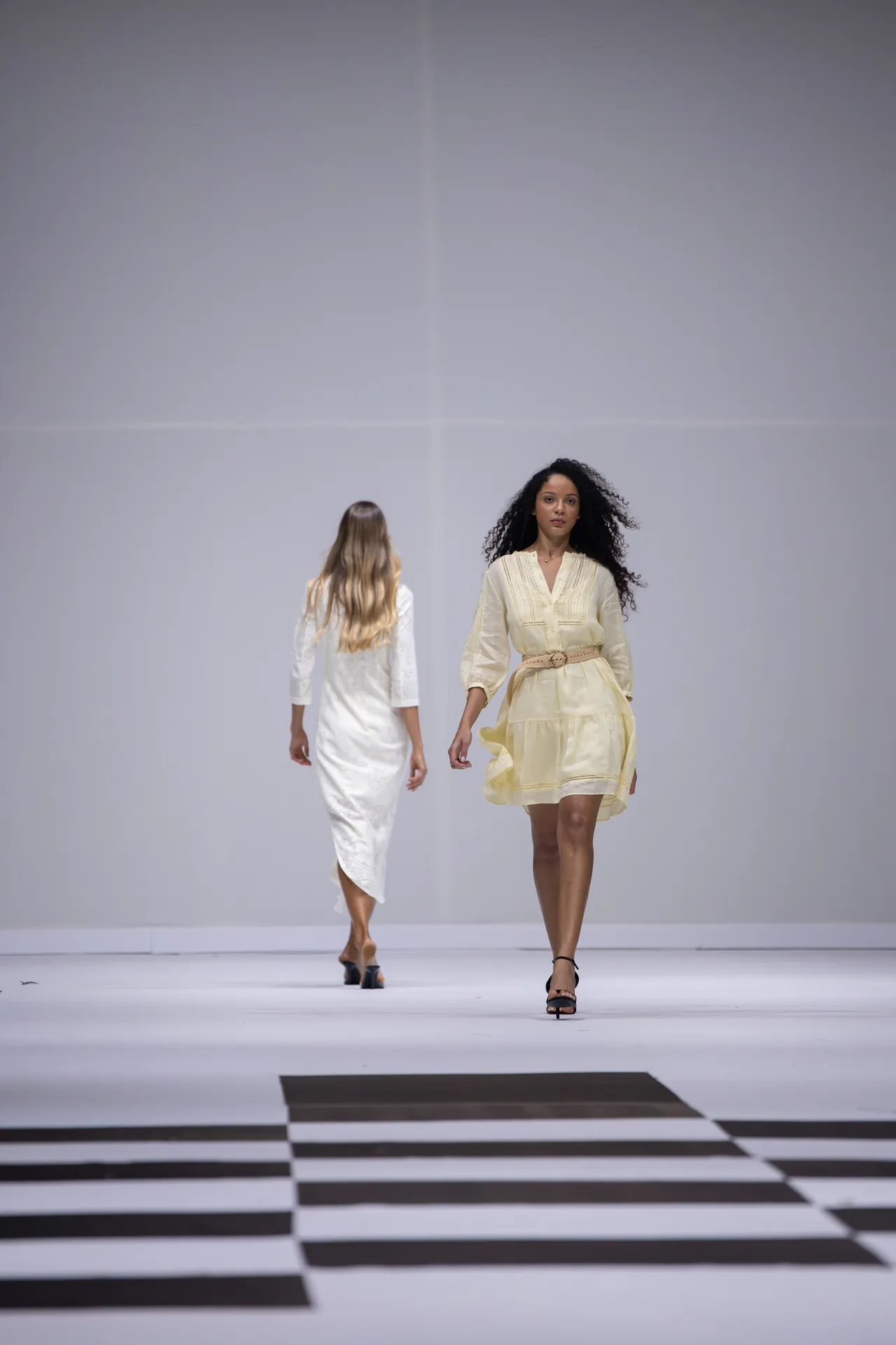 Fashion runway with two models; the foreground model wears a pale yellow dress with a belt and black sandals, walking toward the camera while another model in white walks away behind her.