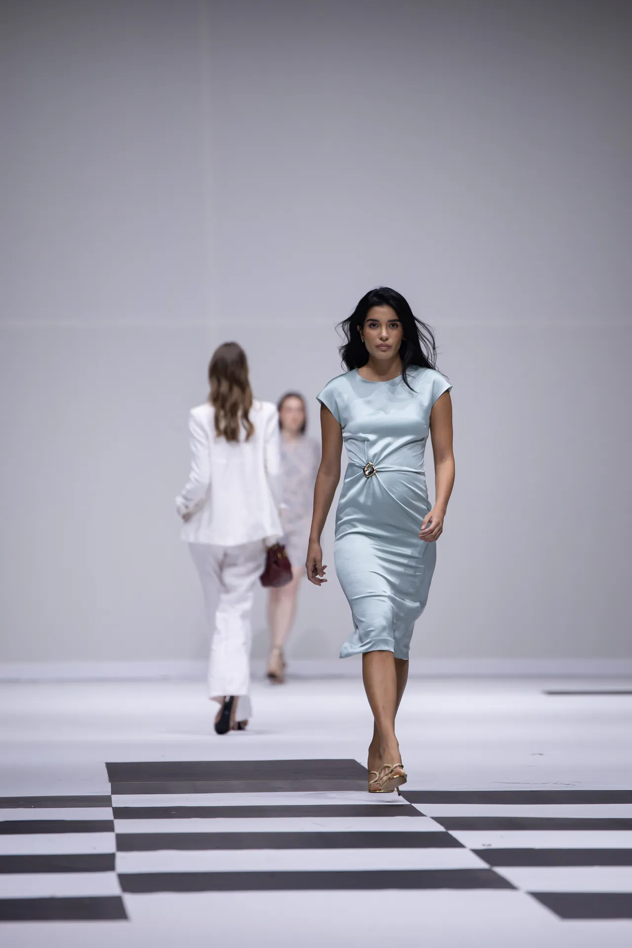 Female model wearing a light blue satin dress walks down a fashion runway with a black-and-white checkered floor and other models in the background.
