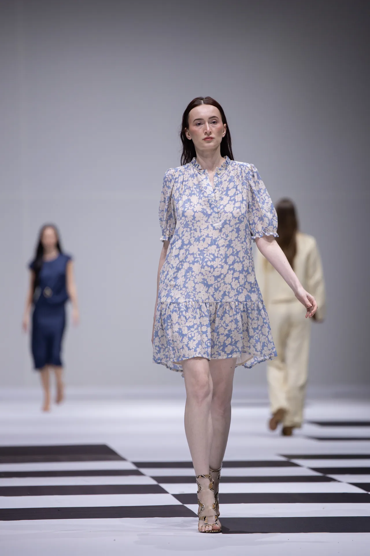Model walking a fashion show in a light blue floral dress on a black-and-white checkered runway.