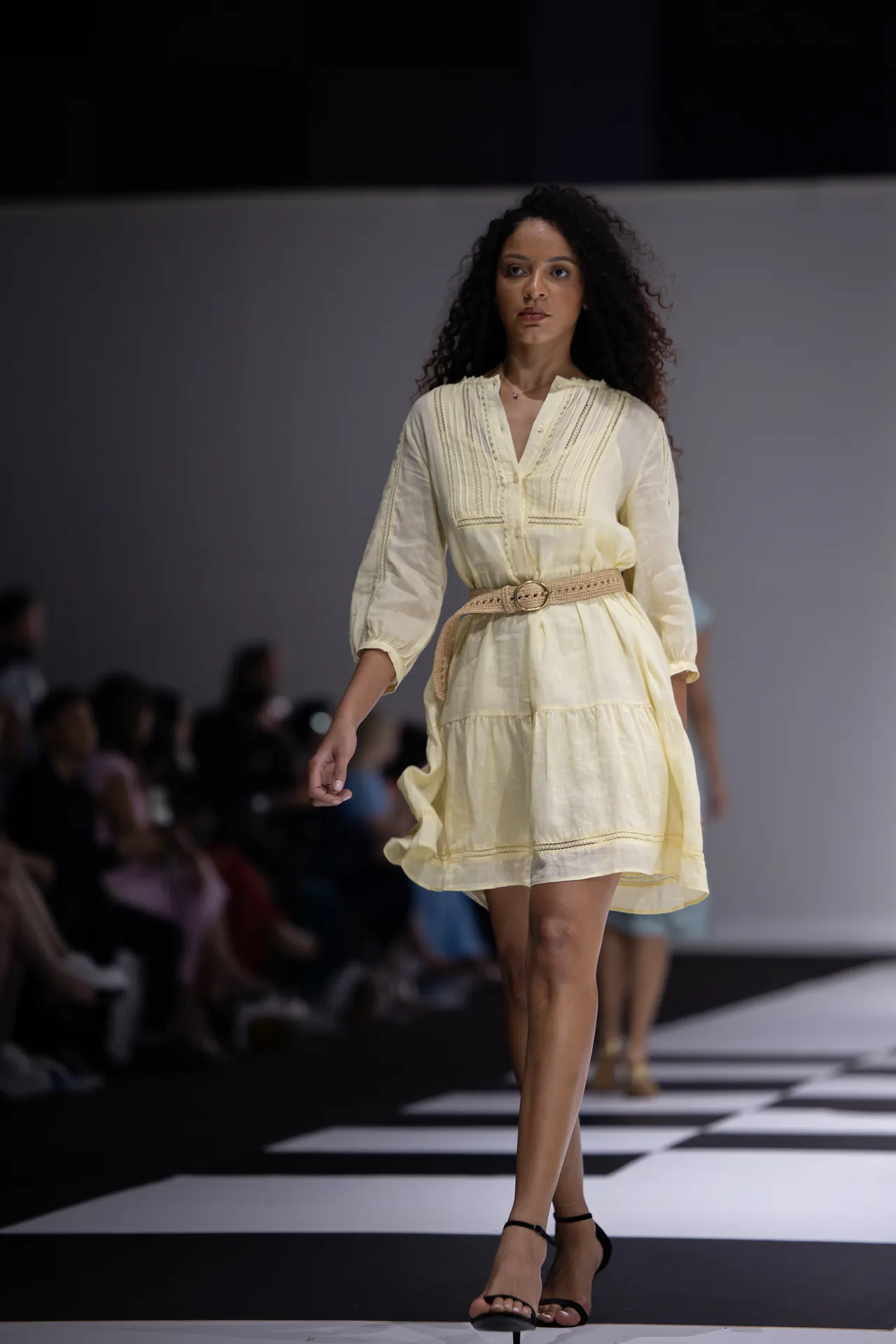 Model walking a fashion runway in a pale yellow dress with embroidered detailing and a beige belt, black sandals, audience in the background.