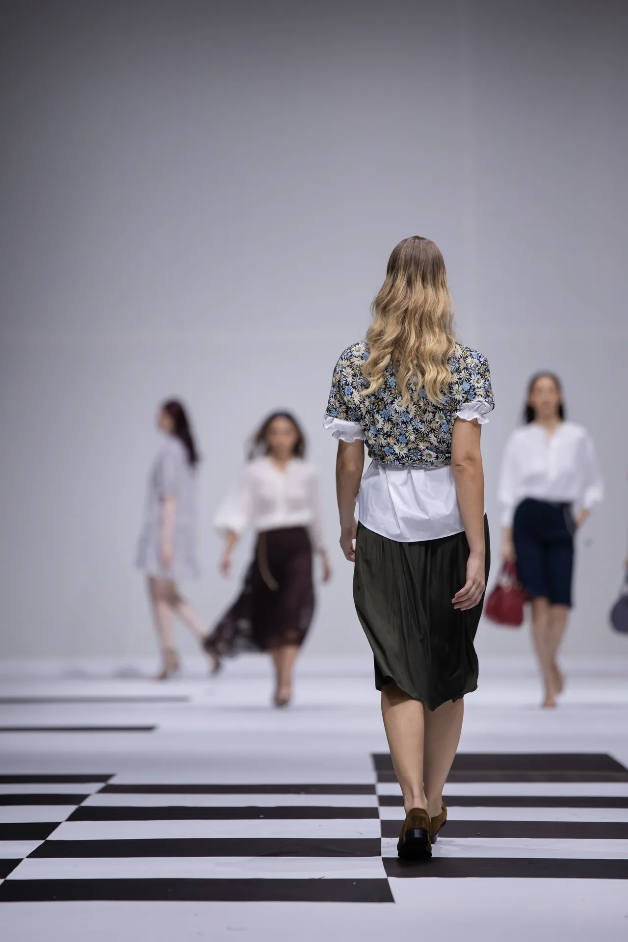 Model walking away on a black-and-white checkered runway, wearing a floral top and white waist-length blouse with a dark skirt; other models blurred in background.